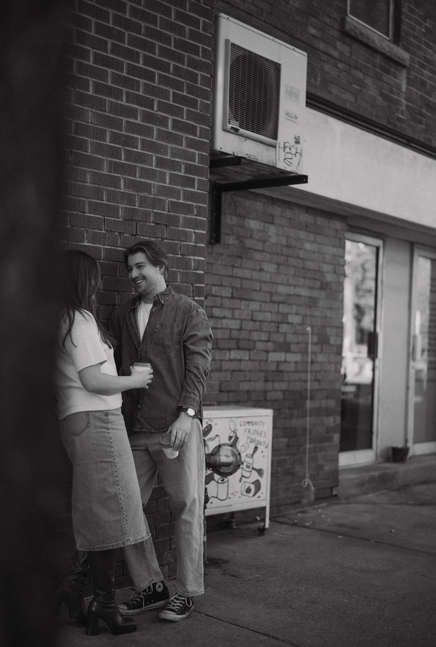 A black and white photo of two people talking while standing against a brick wall outside a building.