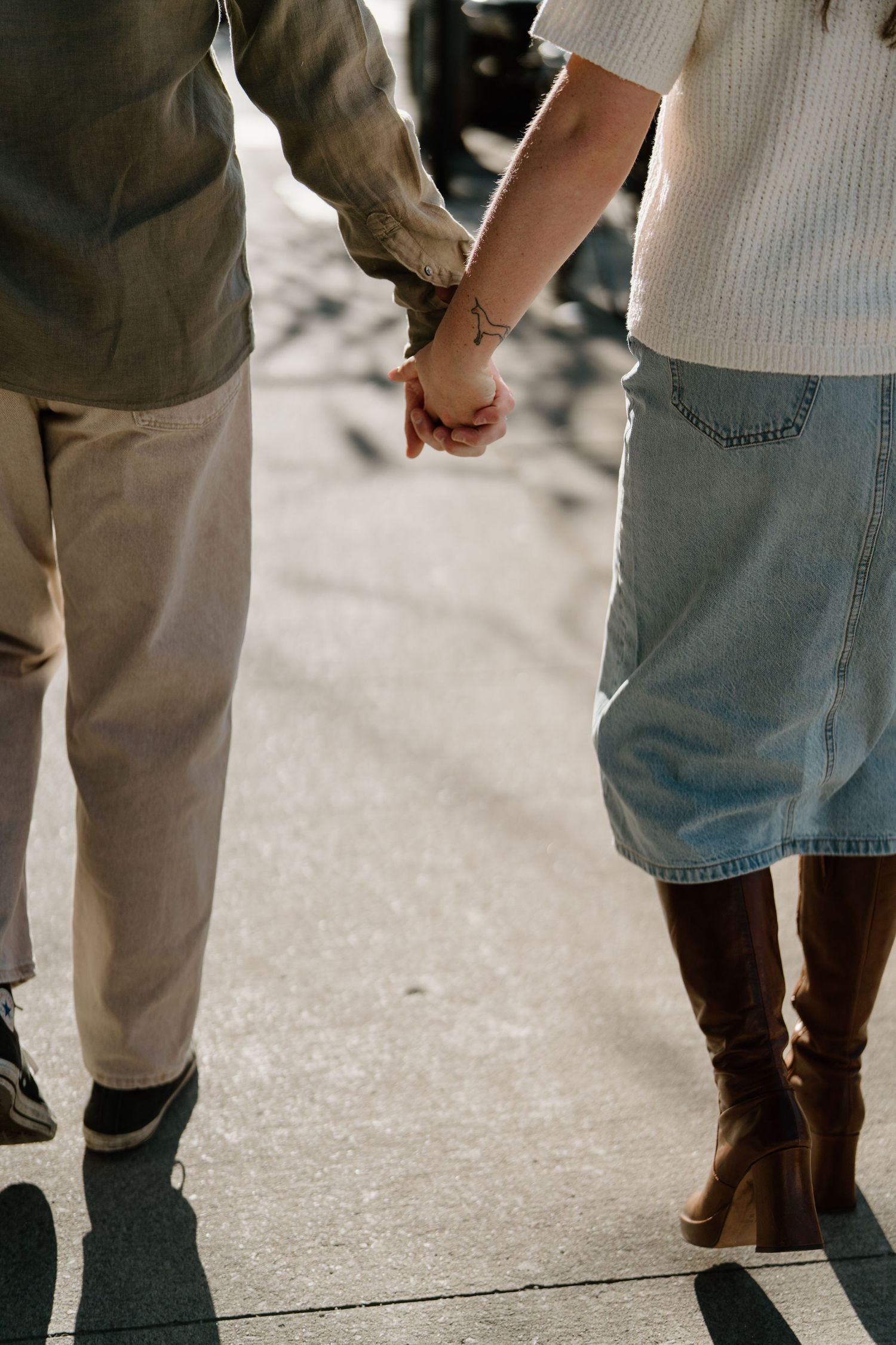 Two people holding hands while walking on concrete, wearing casual clothing with brown boots and khaki pants.