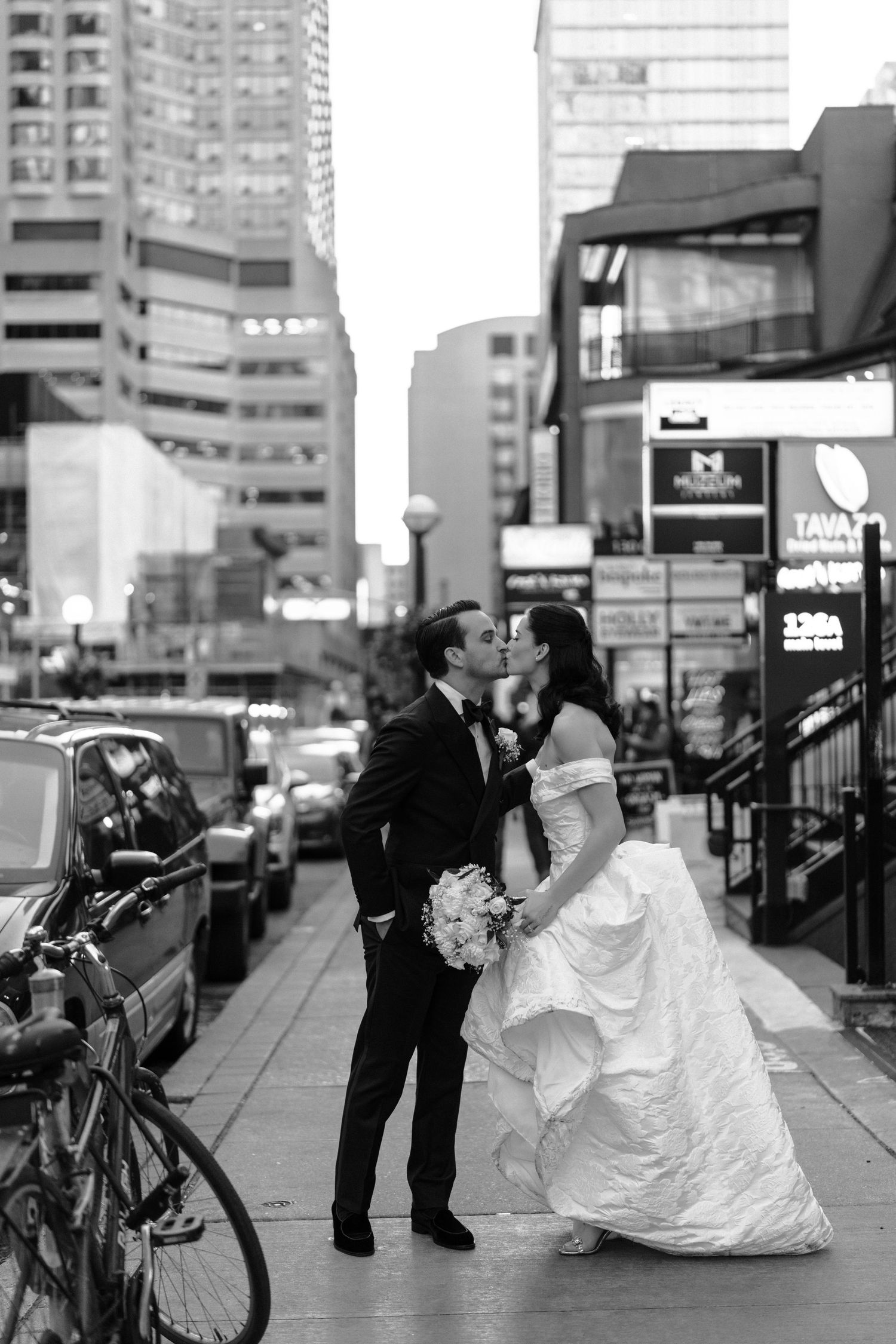 A bride and groom share a romantic moment on a city sidewalk with urban buildings and a bicycle in the background.