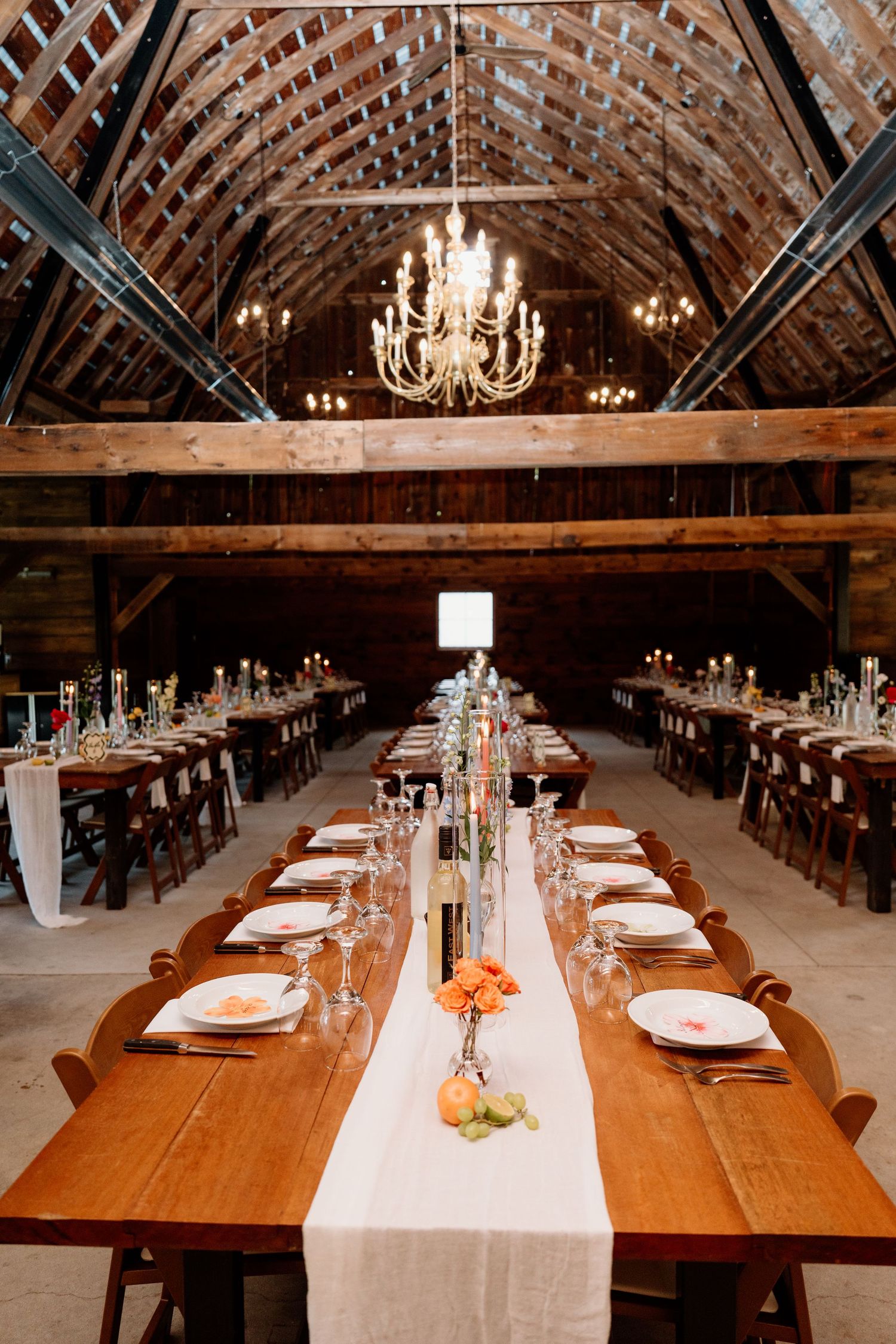 Long wooden tables set for a wedding reception under vaulted ceiling with elegant chandeliers in a rustic barn.