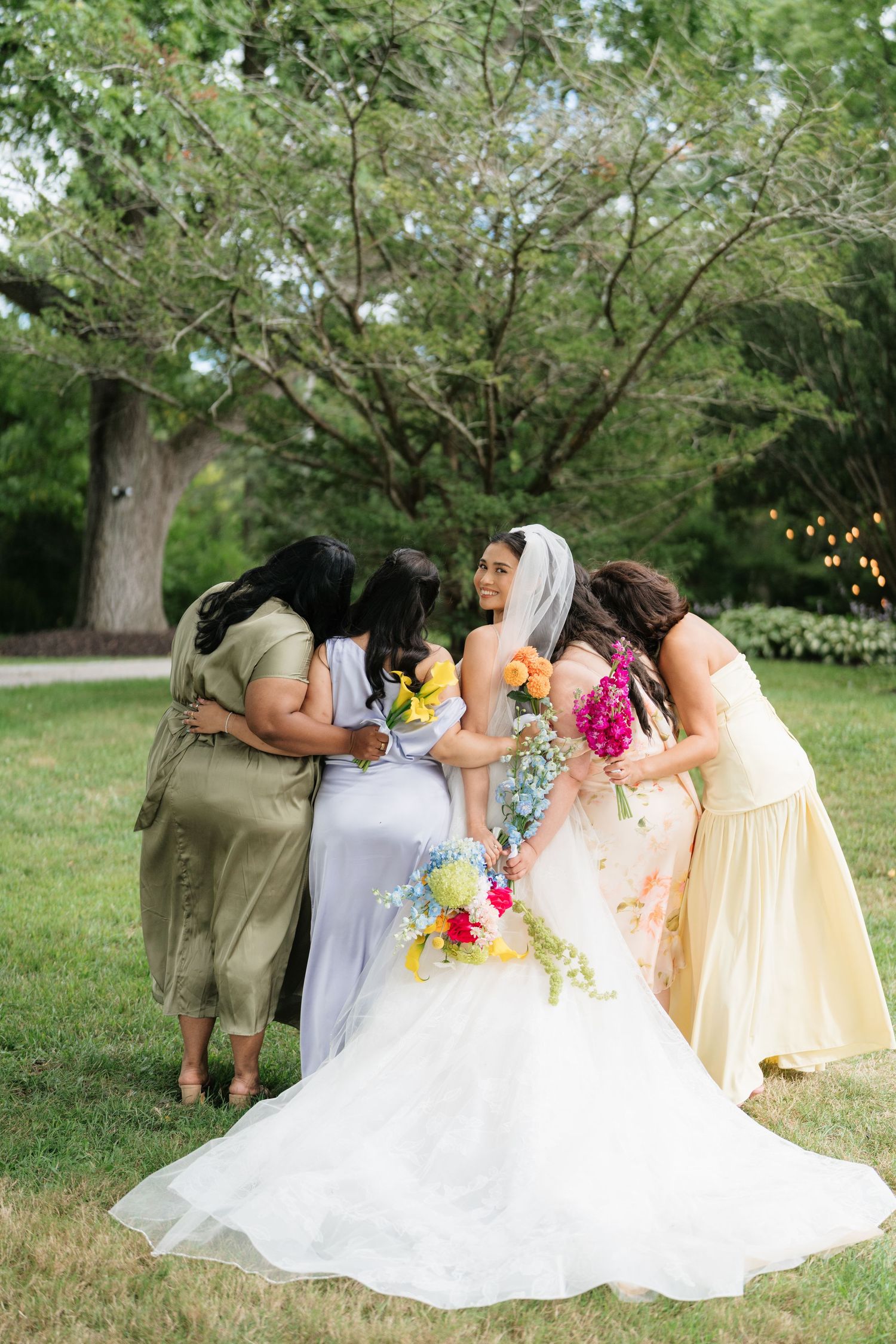 A group of bridesmaids in colorful dresses huddle together with the bride in a white gown on a grassy lawn.