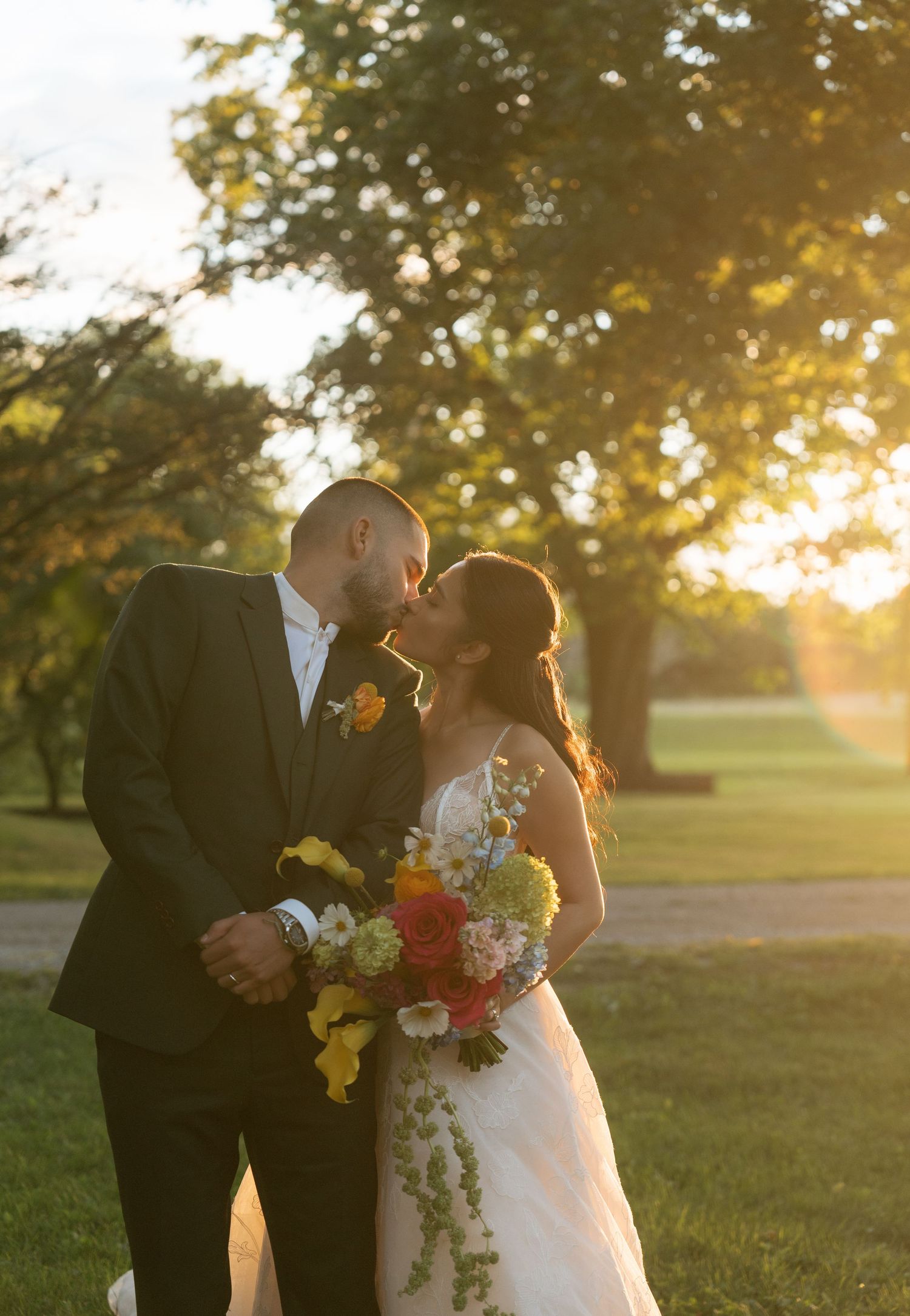 A couple shares a romantic kiss during golden hour sunset in a grassy field with soft sunlight filtering through trees.