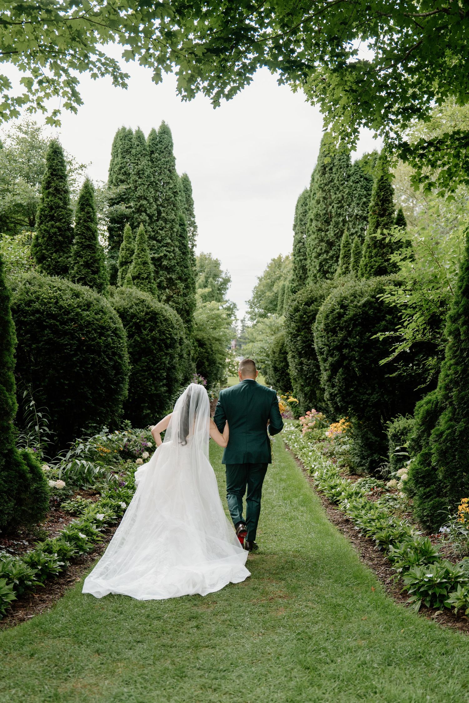 A couple walks down a lush garden path lined with tall evergreen trees and blooming flowers on their wedding day.