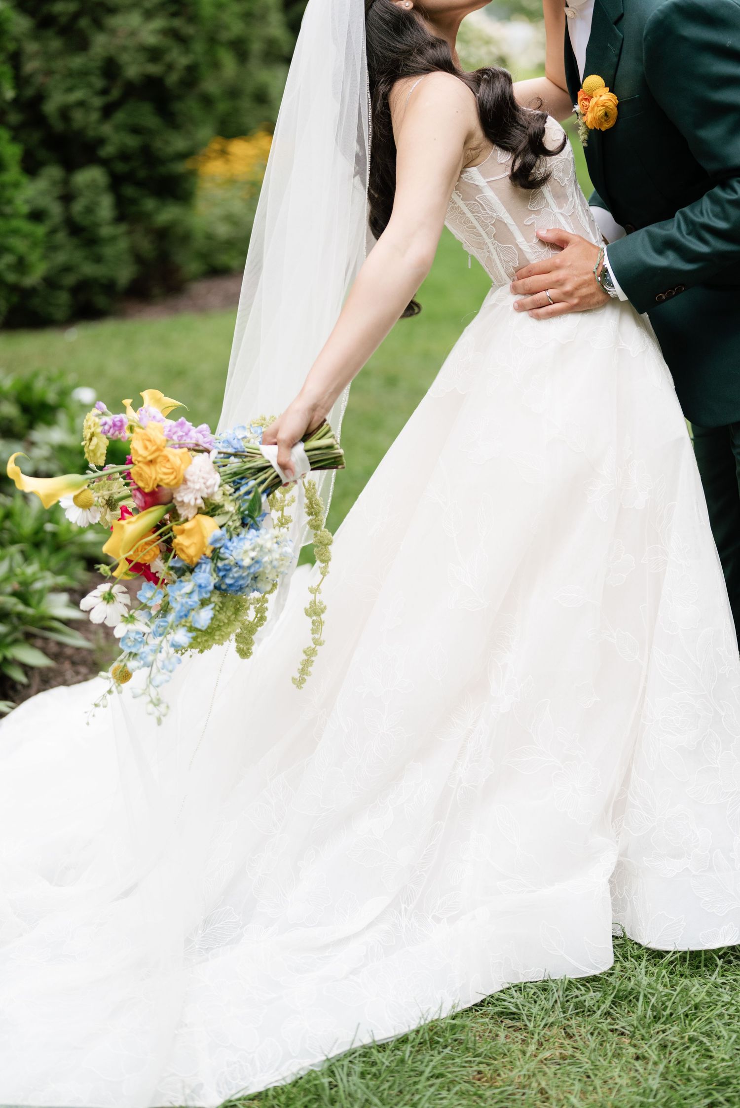 Elegant white wedding dress with tulle skirt flows across green grass while holding a vibrant summer bouquet.