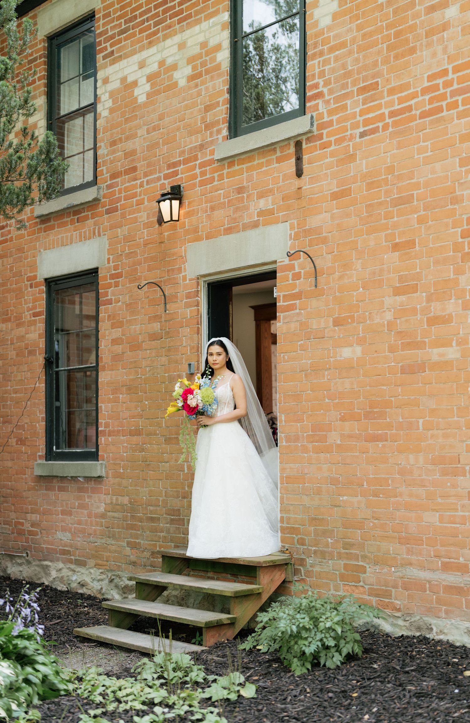 A bride in a white wedding dress stands in the doorway of a historic red brick building holding a colorful bouquet.