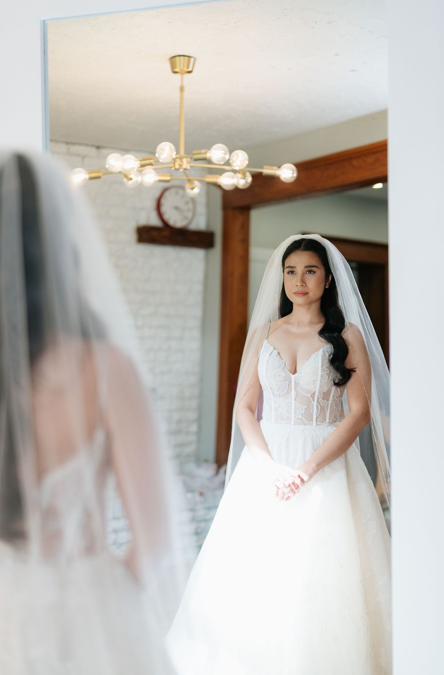 Elegant wedding gown with lace bodice and flowing tulle skirt shown in mirror reflection under modern chandelier.