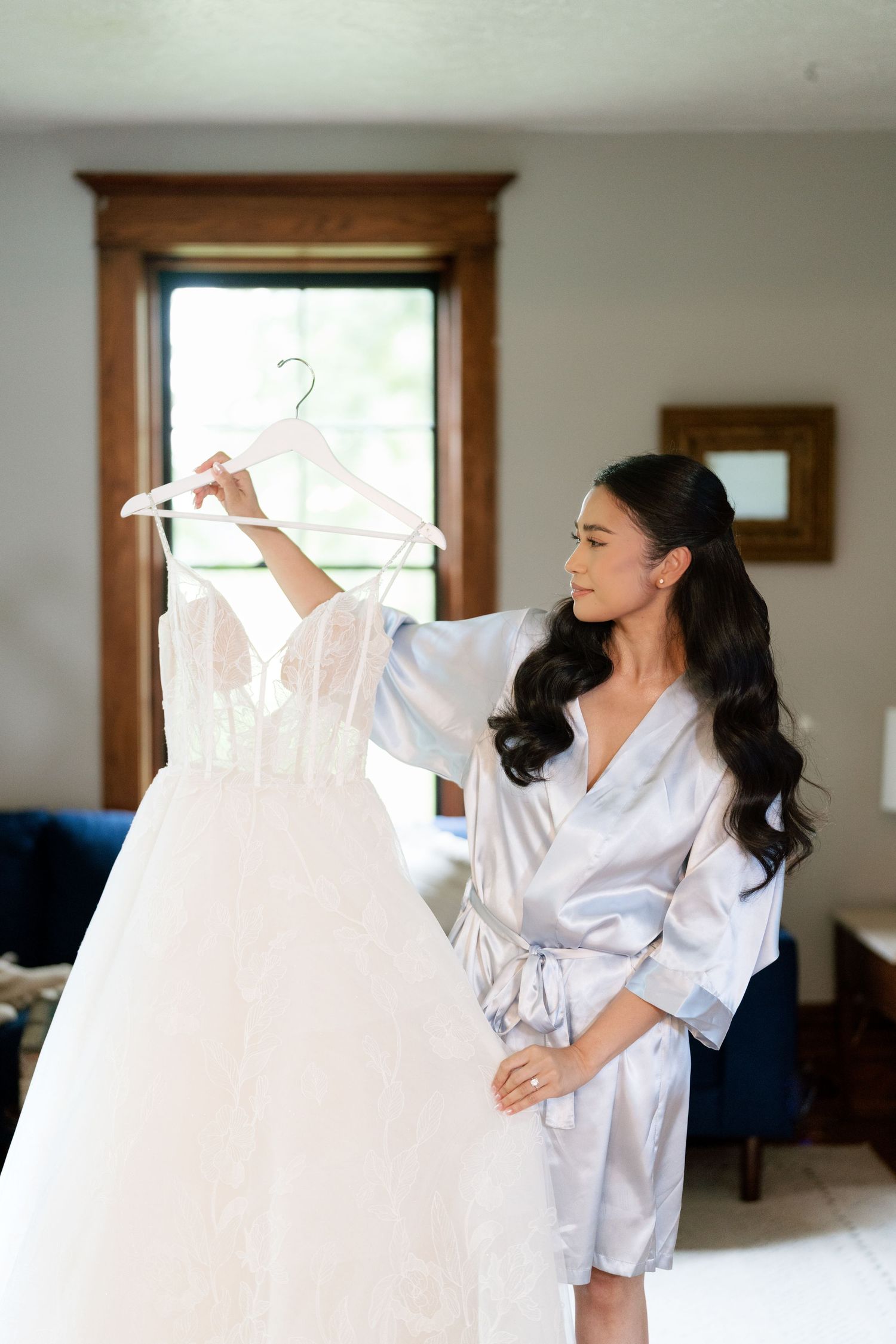 A person in a silk robe examines a white wedding dress on a hanger during wedding preparations.