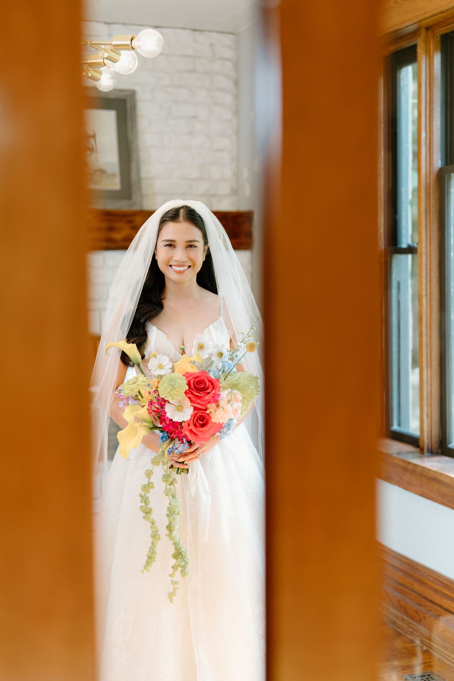 A bride in a white wedding dress and veil holds a vibrant coral and yellow floral bouquet in an indoor setting.
