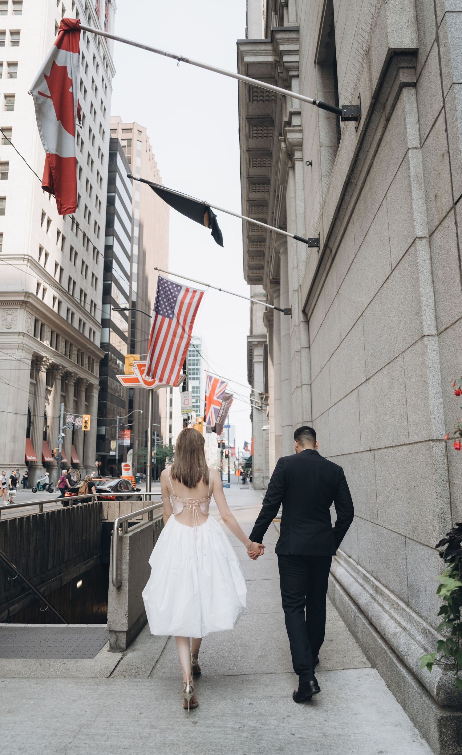 A couple walks hand in hand on a city sidewalk under American and Canadian flags in downtown Toronto.