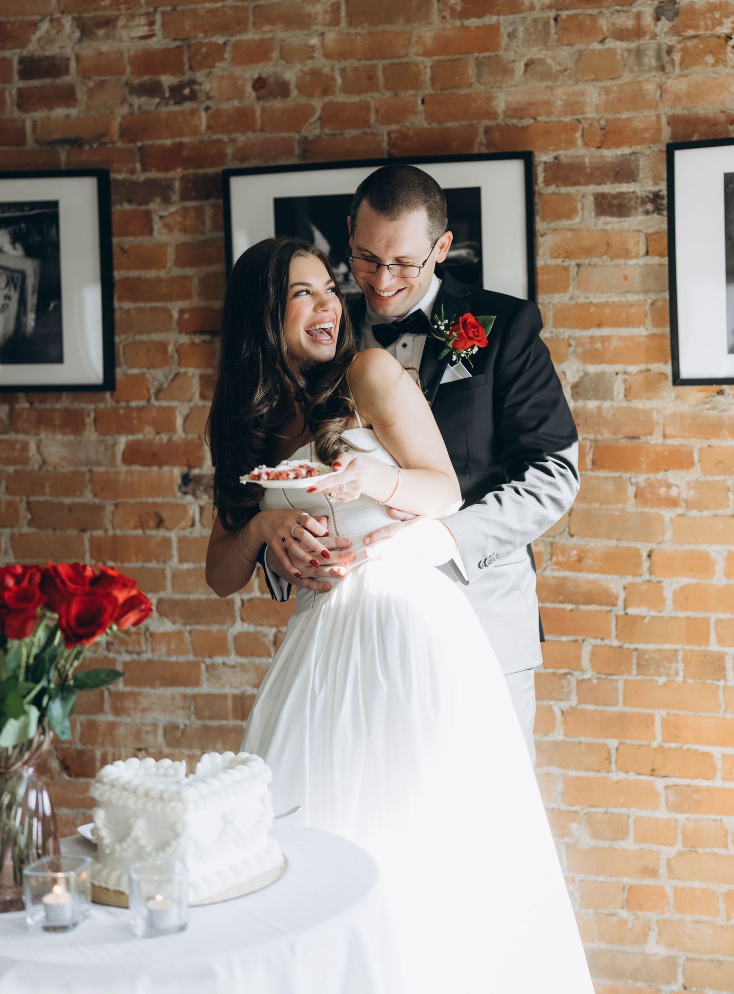Playful wedding moment captured as couple laughs while serving cake, with red roses and brick wall in background.