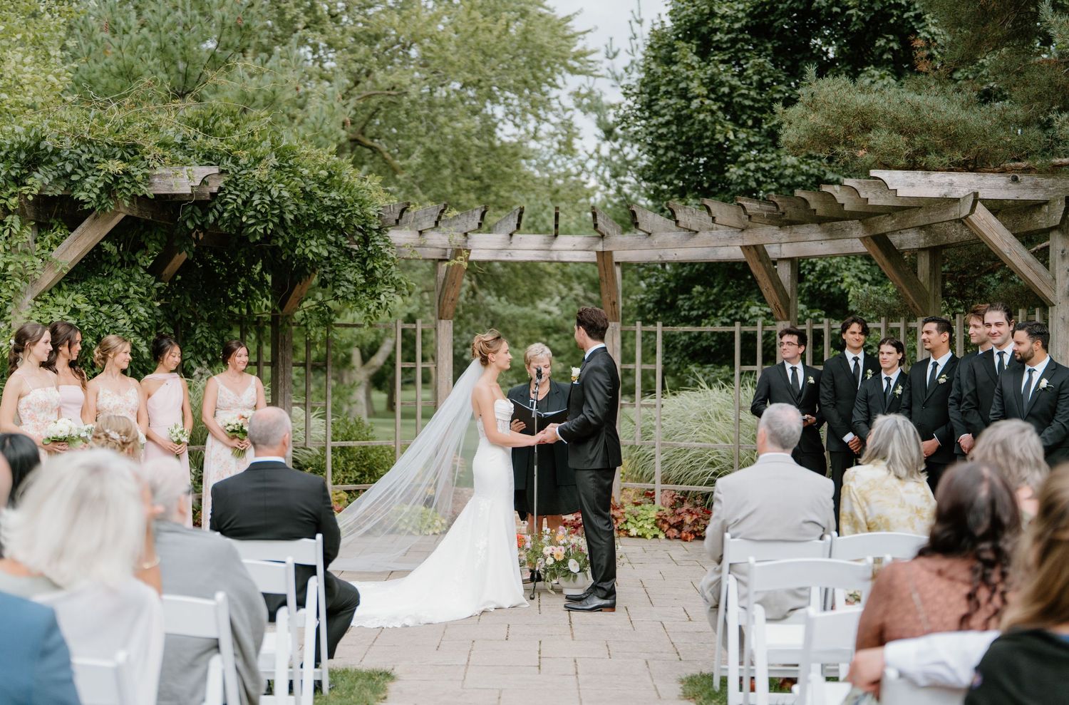 An outdoor wedding ceremony setup with white chairs arranged in rows beneath tall trees and natural greenery.