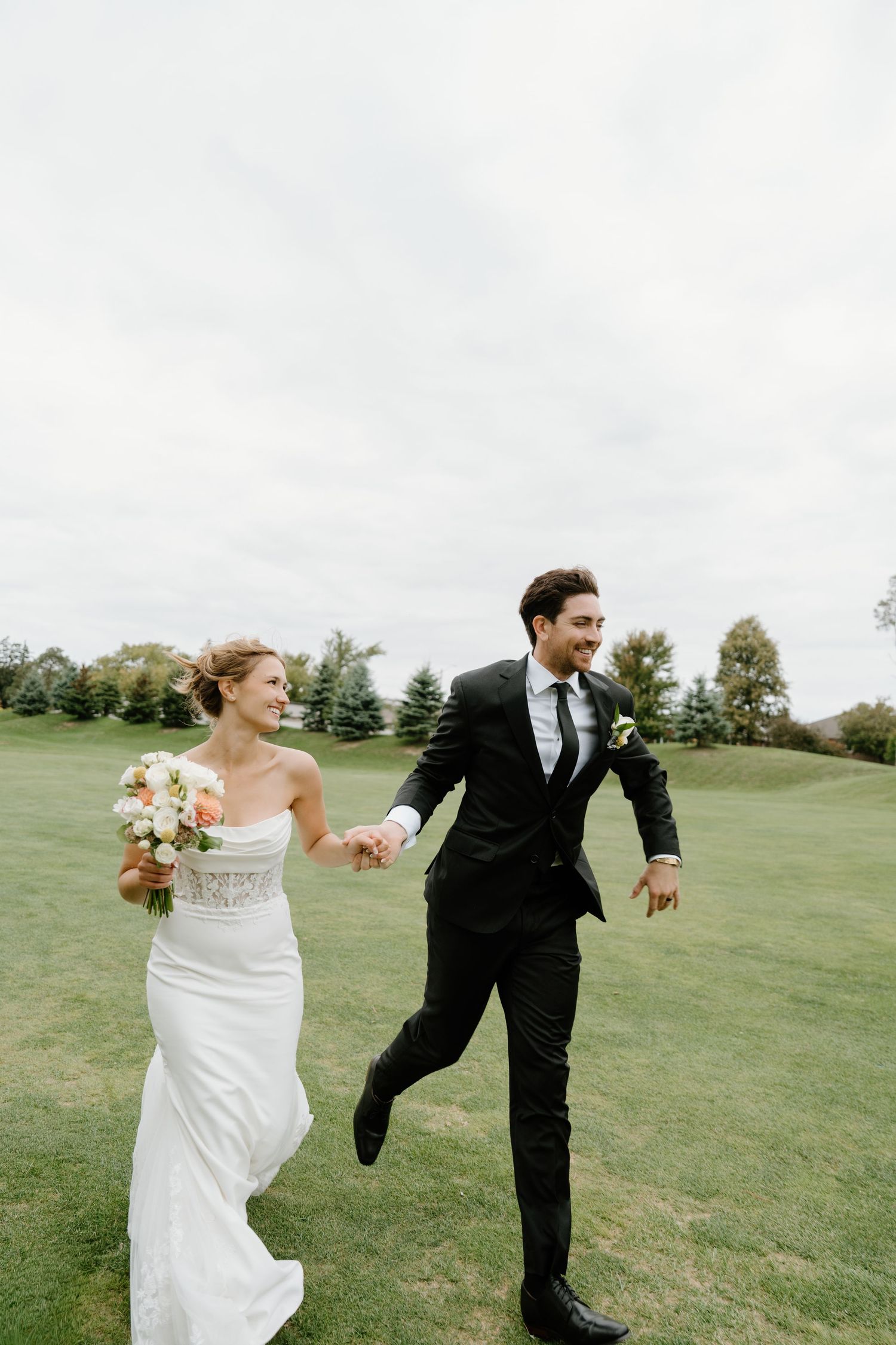 Newlywed couple runs playfully across a green lawn during their wedding photoshoot.
