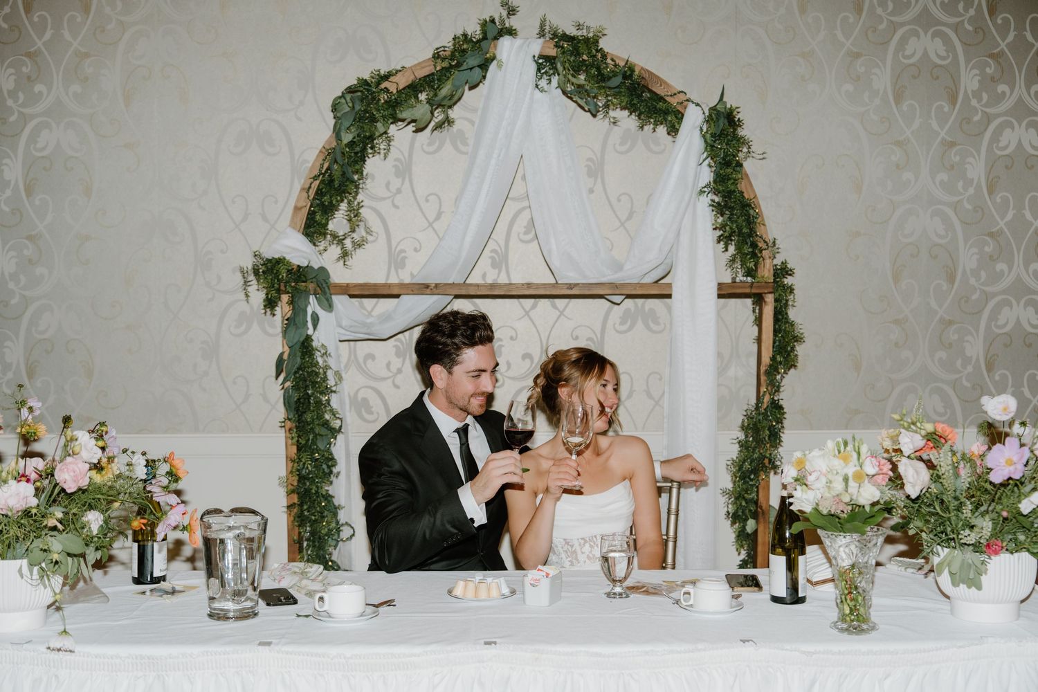 Elegant wedding reception table setting with greenery garland and white linens in a romantic indoor venue.