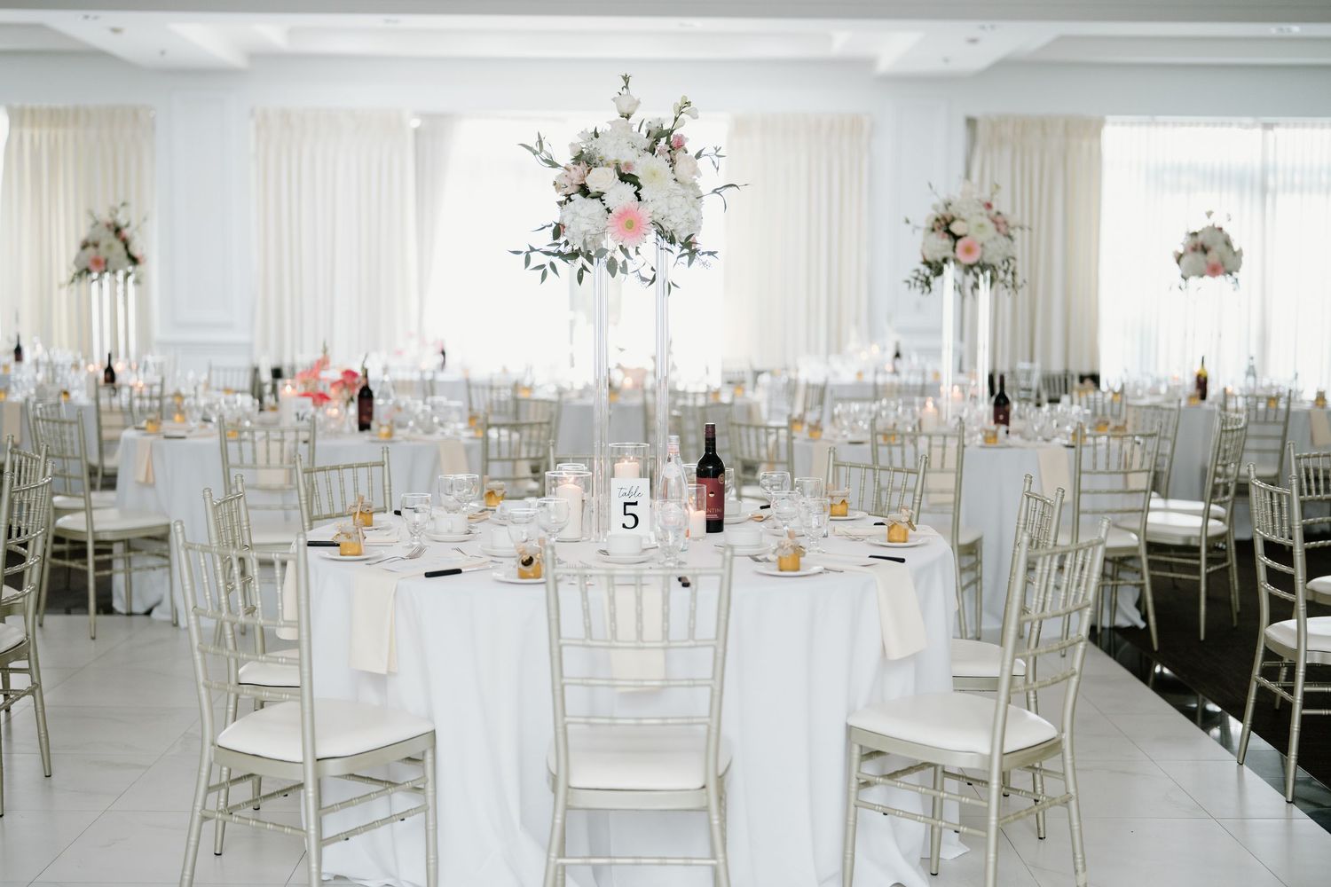 Elegant white wedding reception venue with chiavari chairs and floral centerpieces in a bright, airy space.