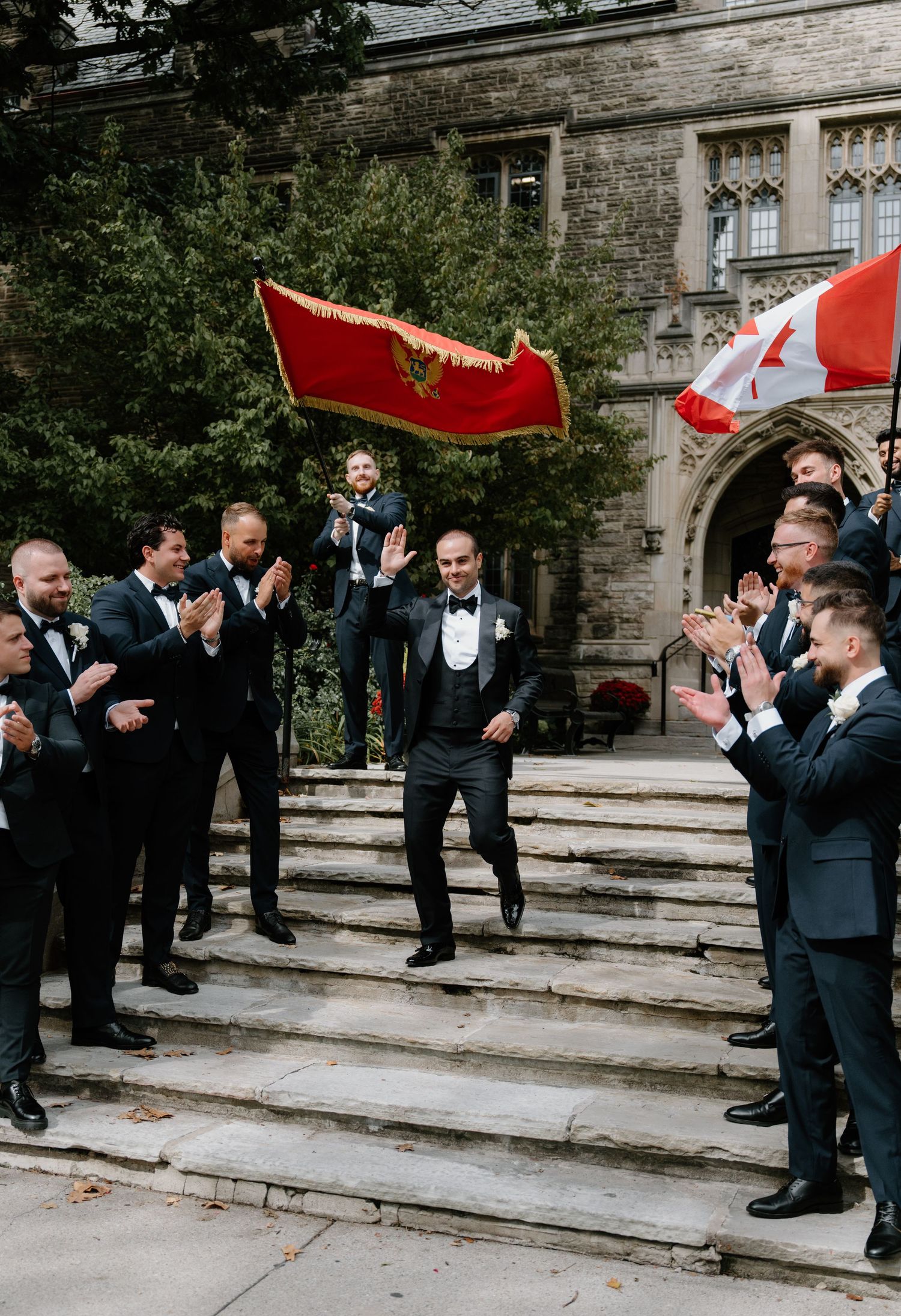 Wedding party groomsmen in black tuxedos pose playfully on stone steps of historic building for fun group photo.