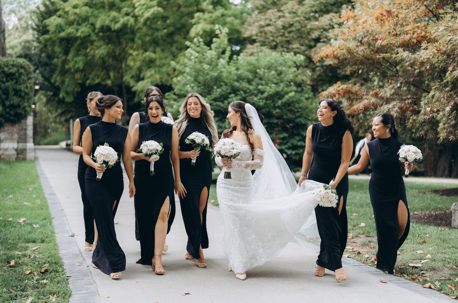 Wedding party in black dresses helps lift bride's white gown while walking through a garden path.