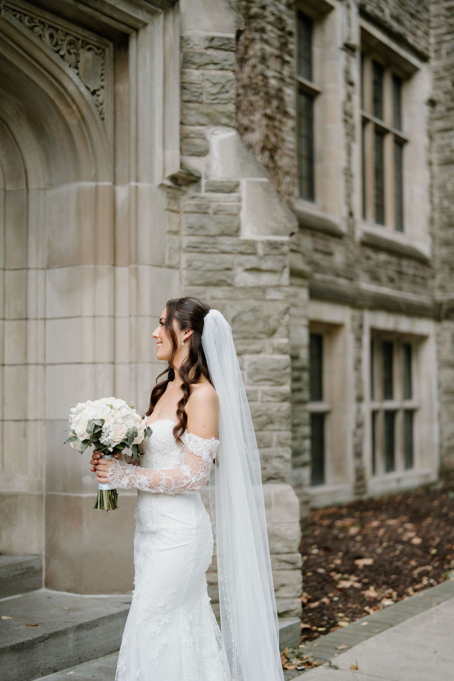 Bride in a white lace wedding dress and long veil poses against a stone building wall.