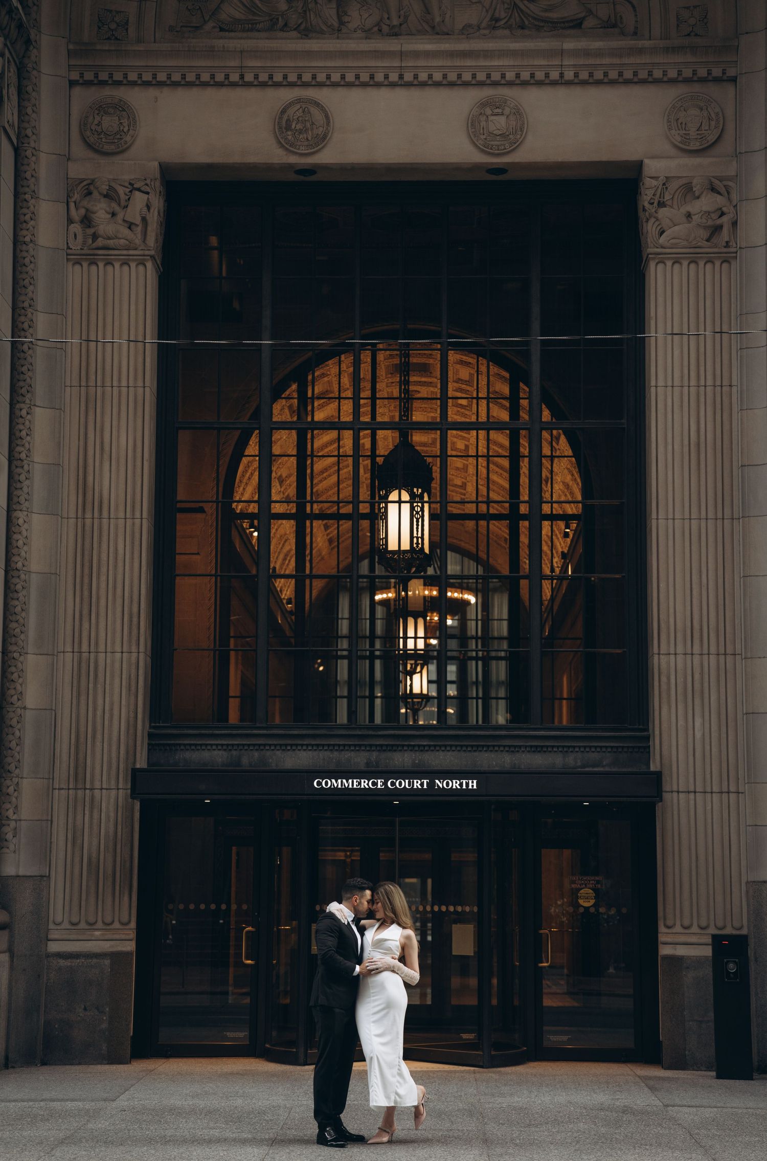 A couple poses in front of an ornate building entrance with classical columns and arched windows.
