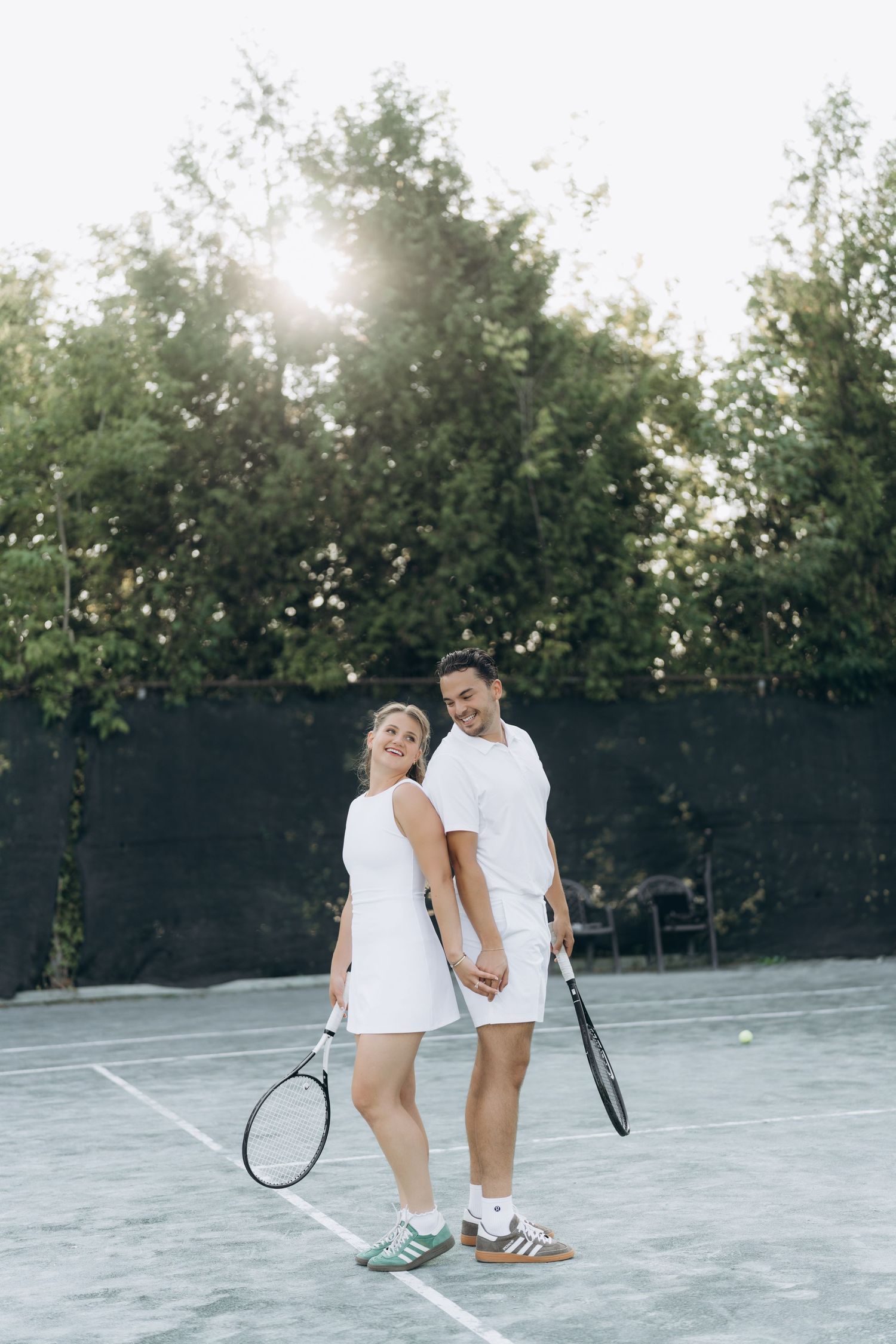 A couple dressed in white athletic wear shares a romantic moment while holding tennis rackets on the court.