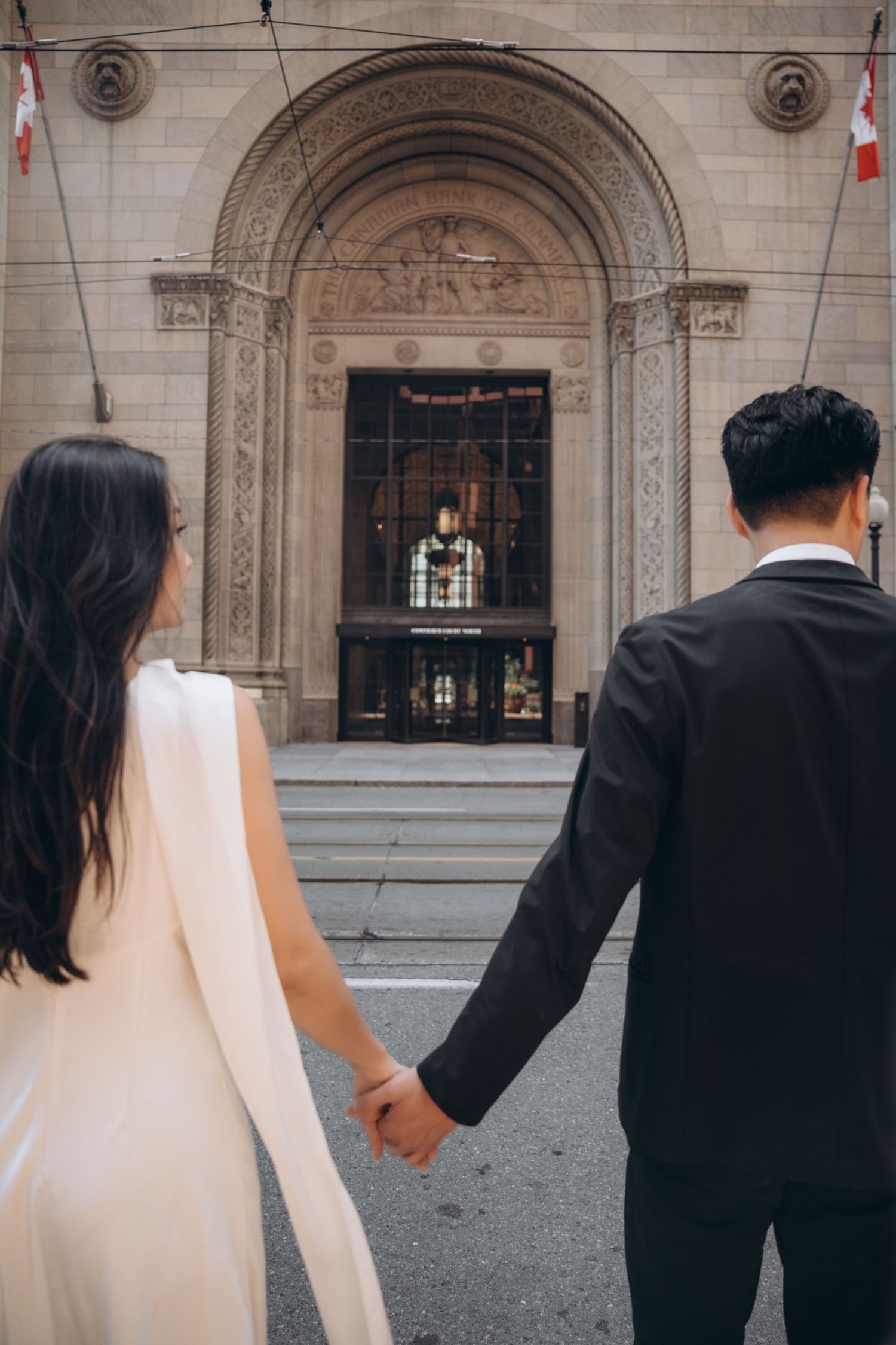 A row of figures in wedding attire holding hands walking in front of an ornate stone building.