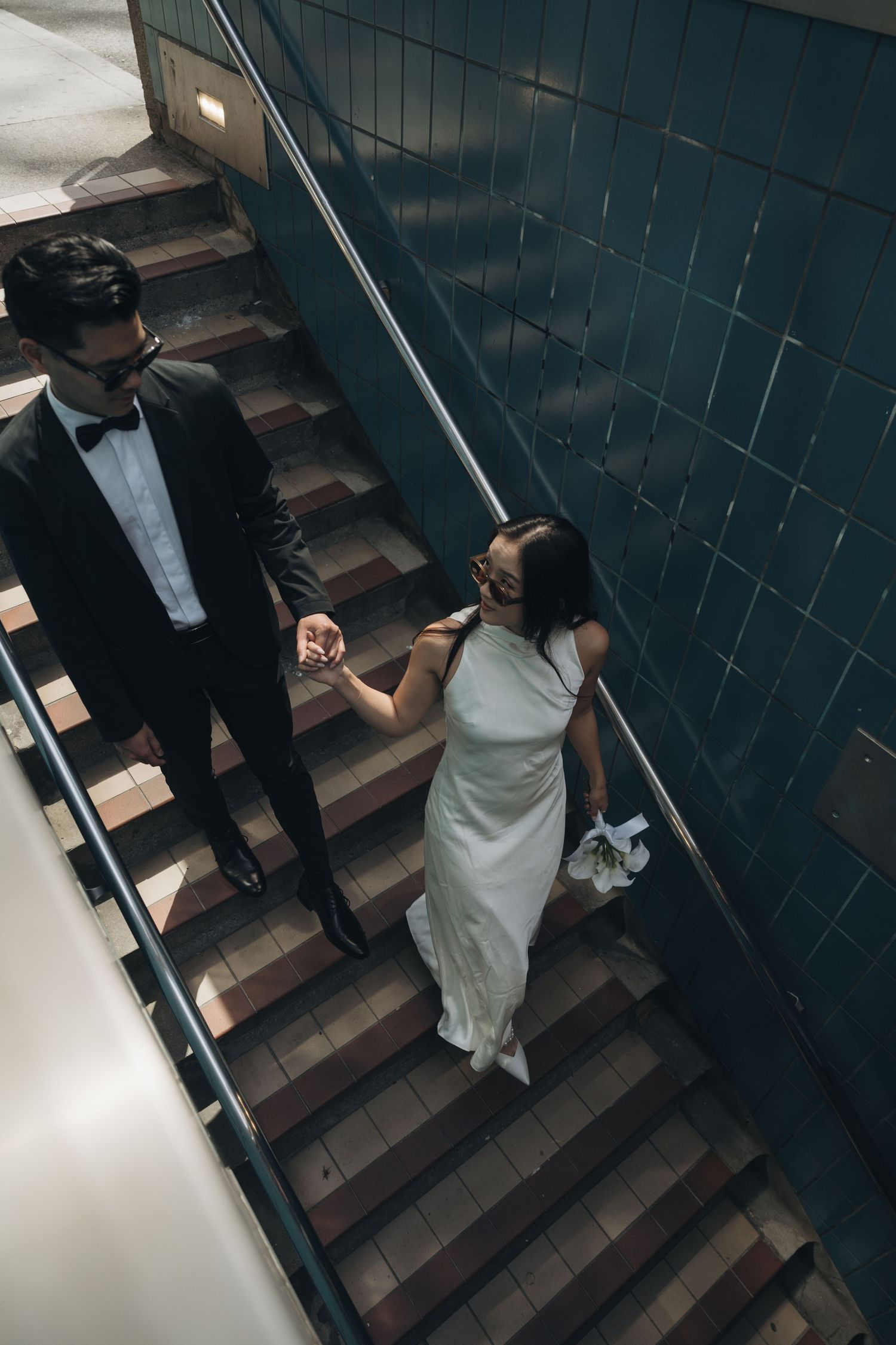 A couple in formal attire holds hands while descending dark stairs in a moody indoor setting.