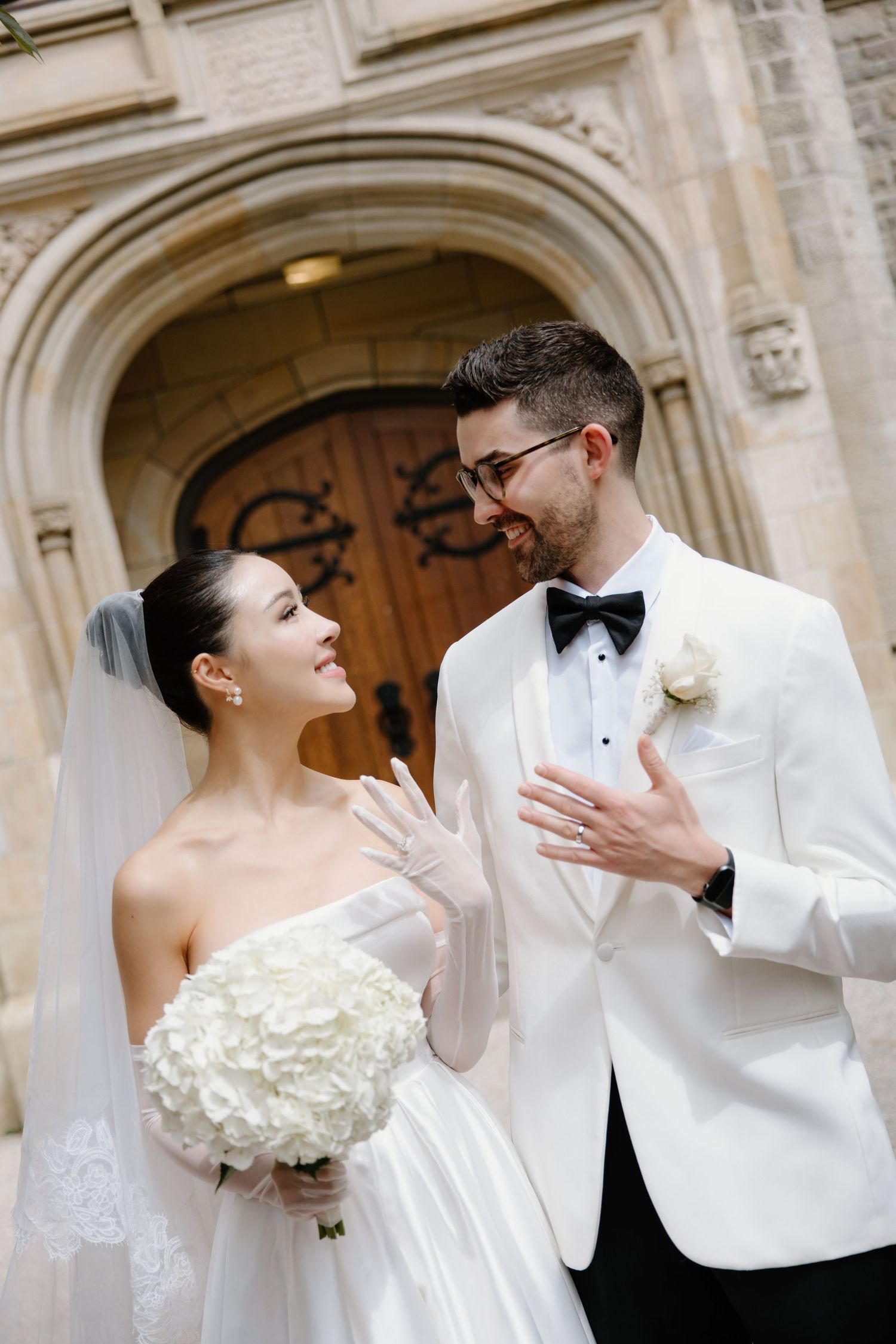 A couple in formal wedding attire share a joyful moment outside an ornate arched doorway with stone architecture.
