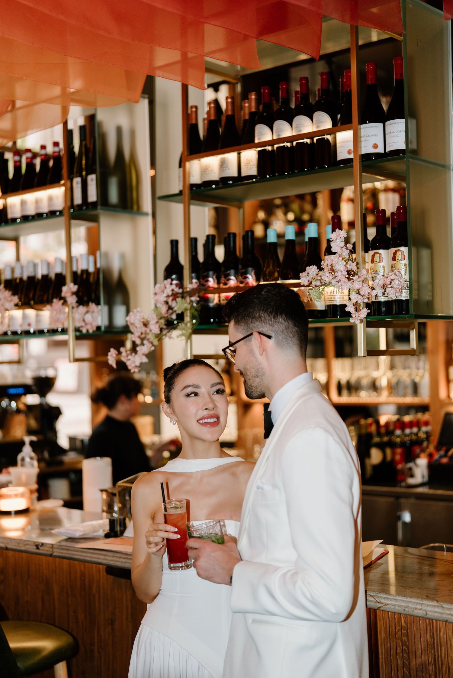 Couple in formal attire shares cocktails at an elegant bar with shelves of bottles and warm lighting behind them.