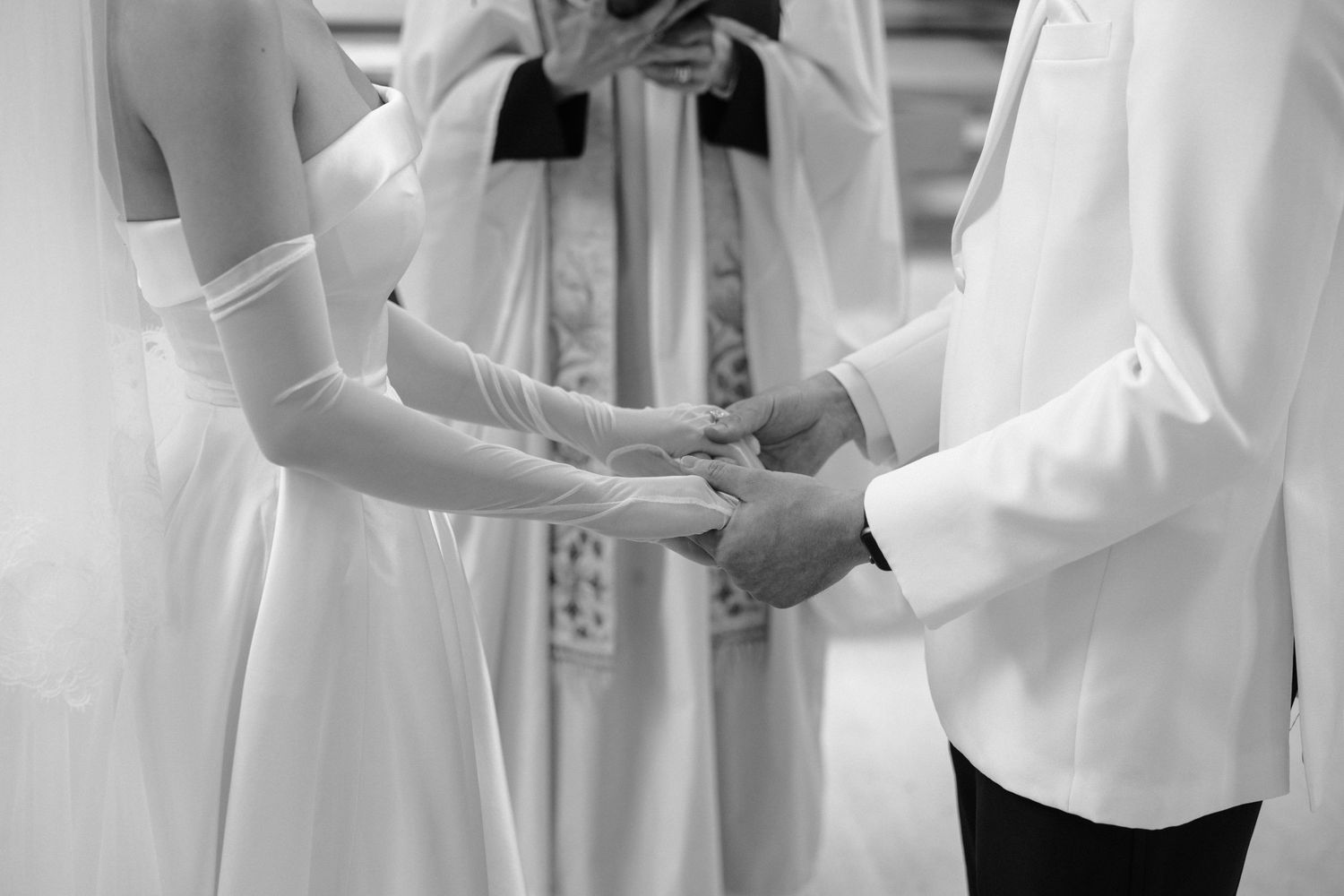 A black and white photograph of hands being joined during a wedding ceremony with an officiant in the background.