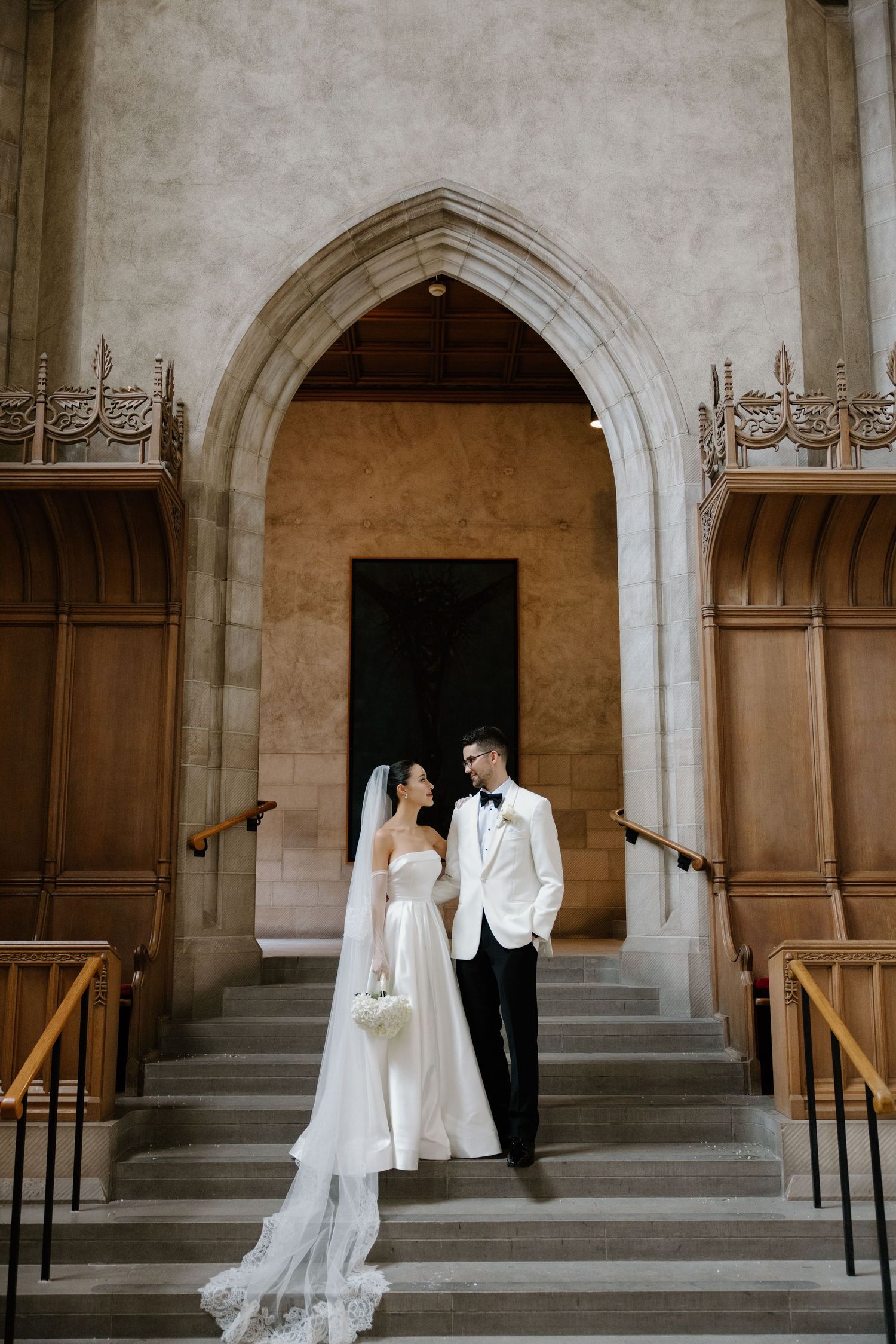 A wedding couple stands in a grand church doorway with Gothic arched entrance and ornate wooden details.