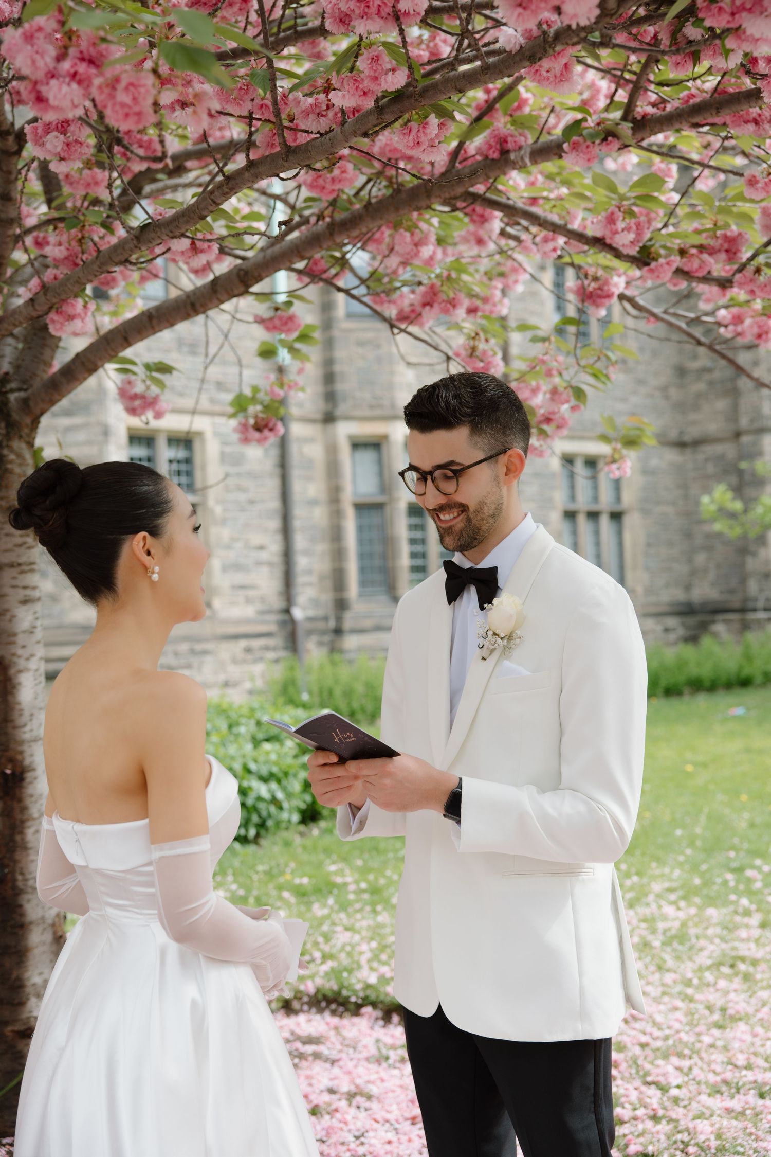 Couple exchanges vows under blooming cherry blossom tree with pink petals falling on spring day.
