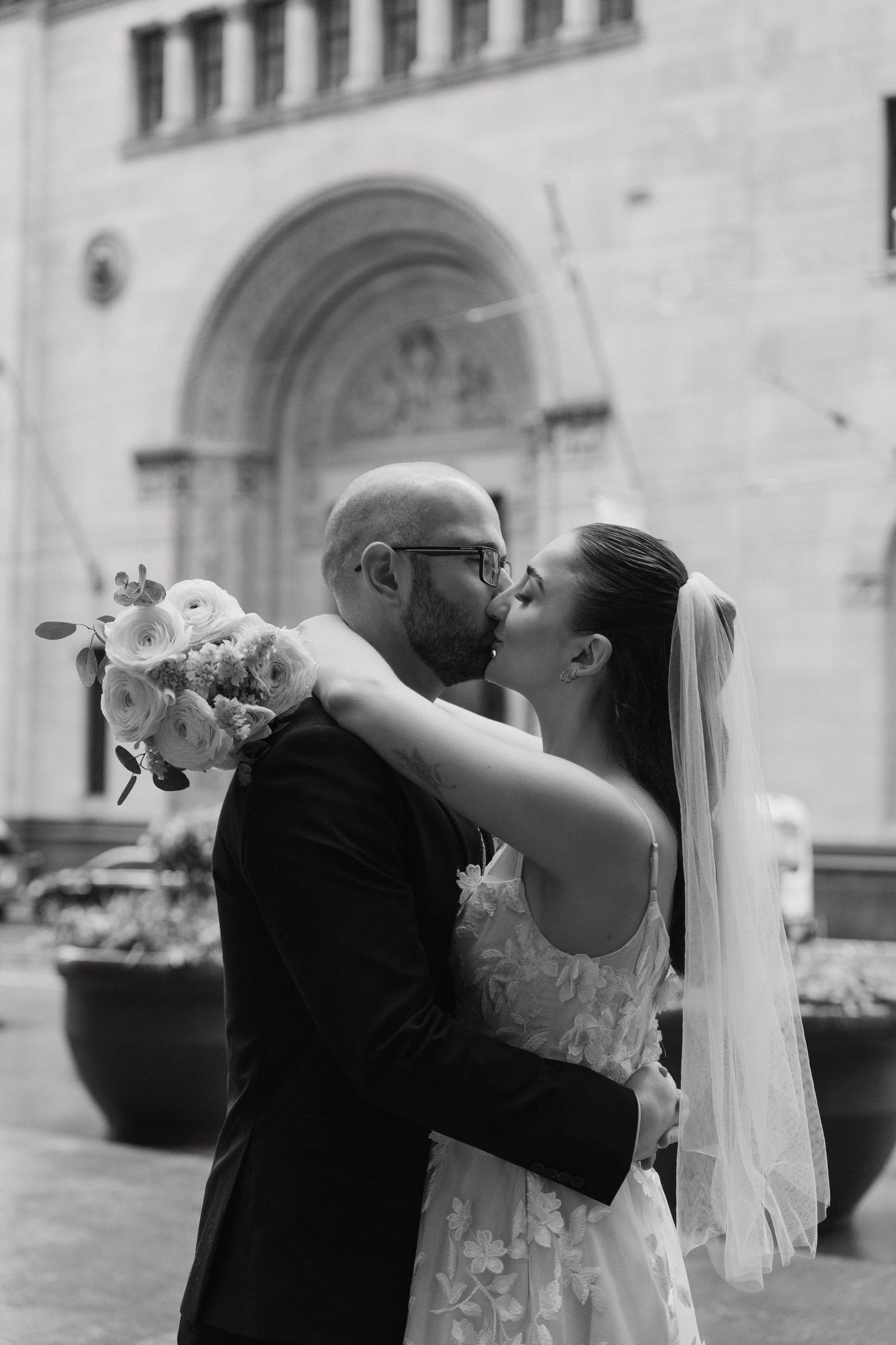 A romantic wedding moment captured in black and white with the couple sharing a kiss against a stone building backdrop.