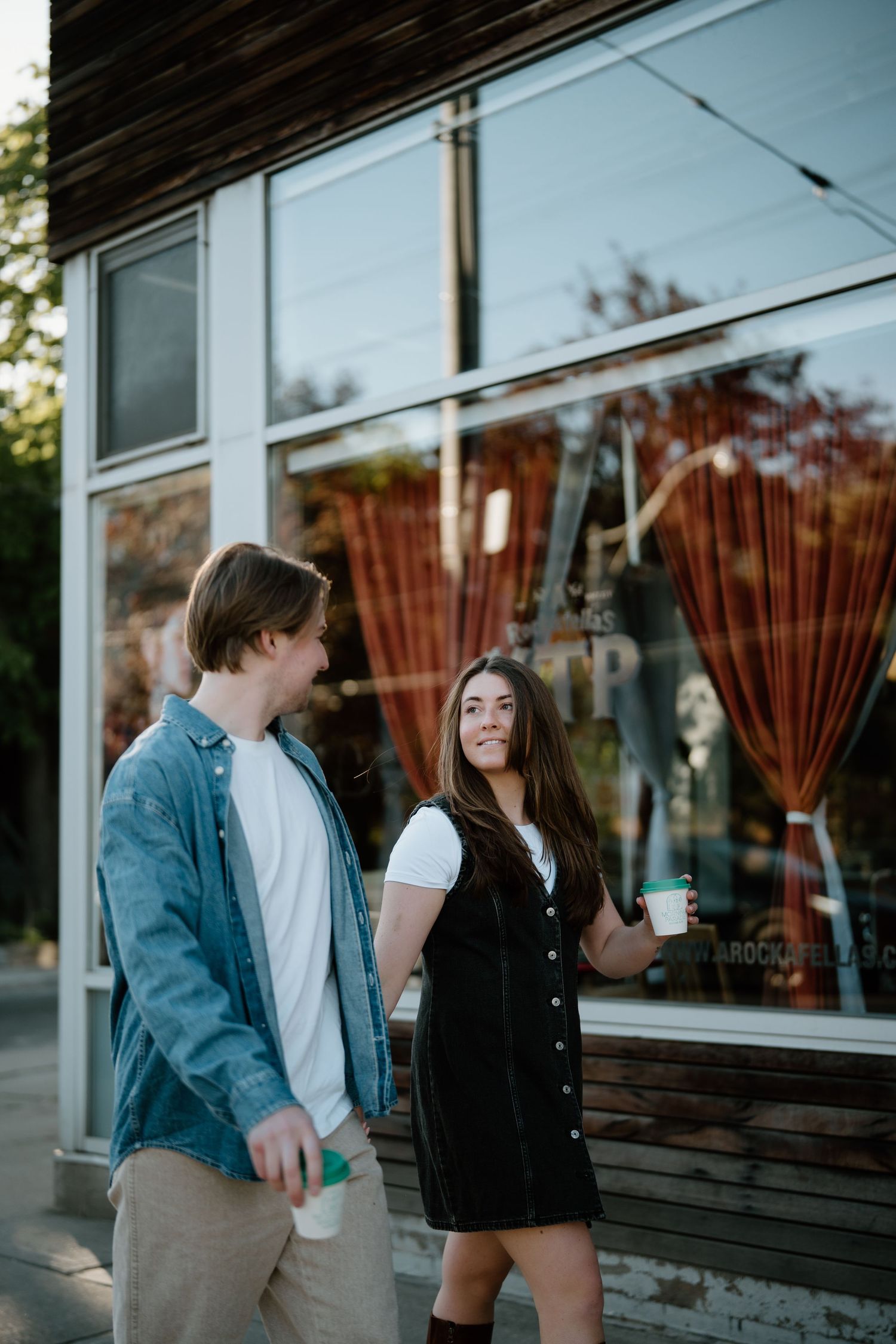 A young couple stands outside a coffee shop window with drinks in hand during golden hour.