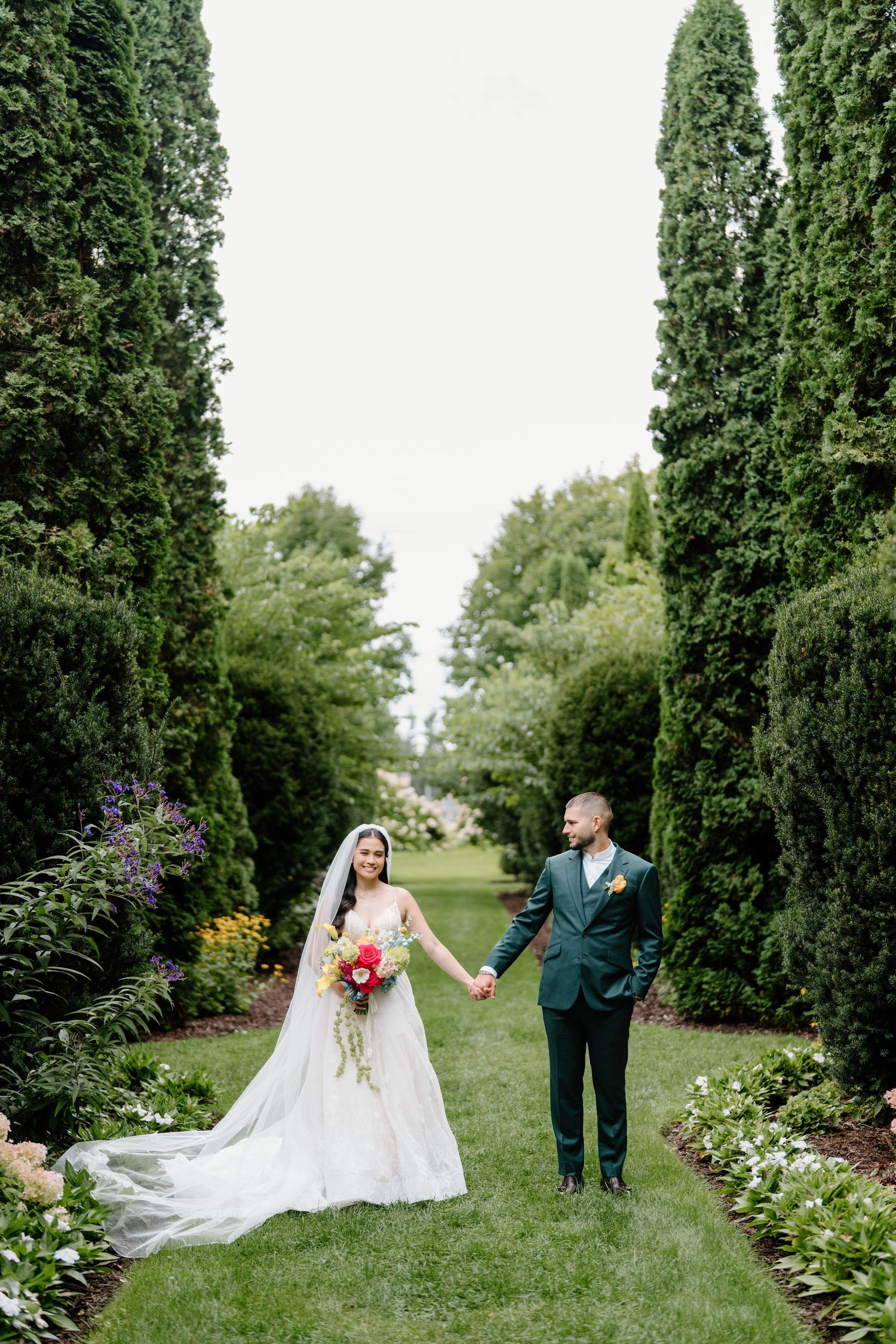 Wedding couple poses in beautiful garden setting with tall cypress trees and blooming flowers.