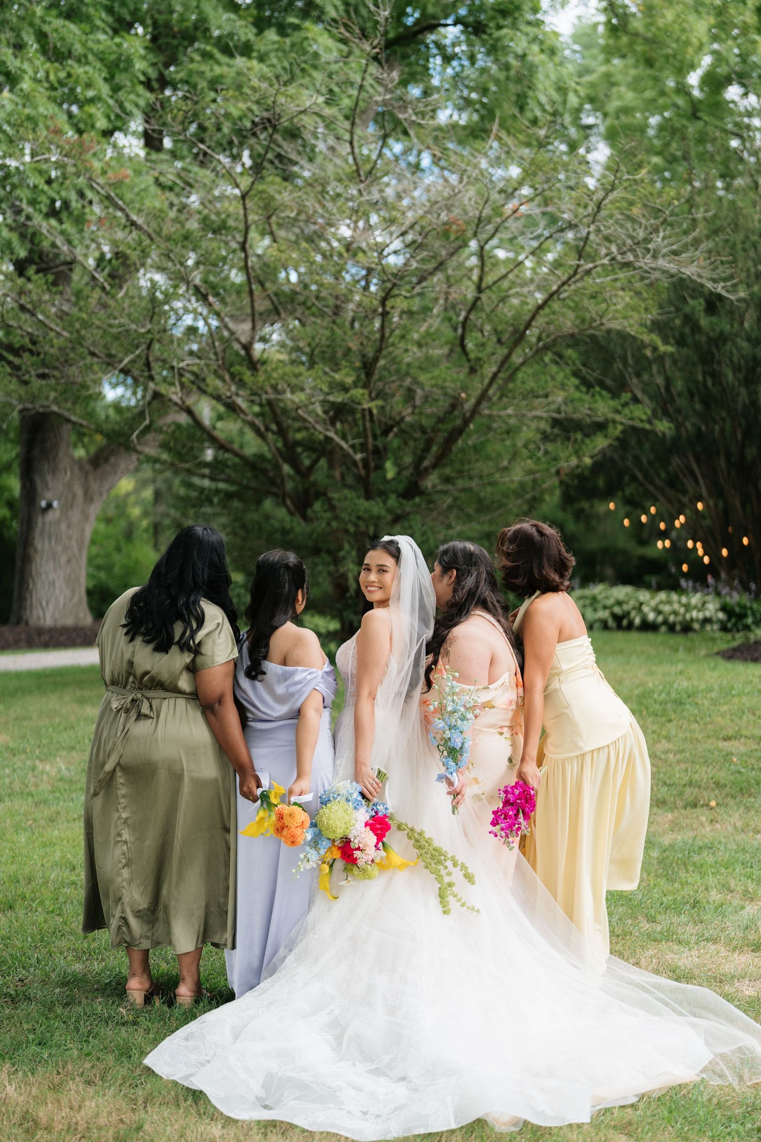 A bride in a white wedding dress stands with bridesmaids wearing pastel dresses in a garden setting with string lights.
