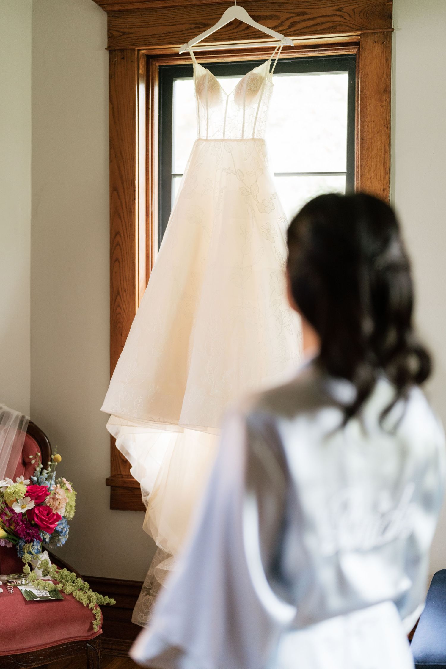 A white wedding dress hangs in a window while bridesmaids in white robes prepare in the background.