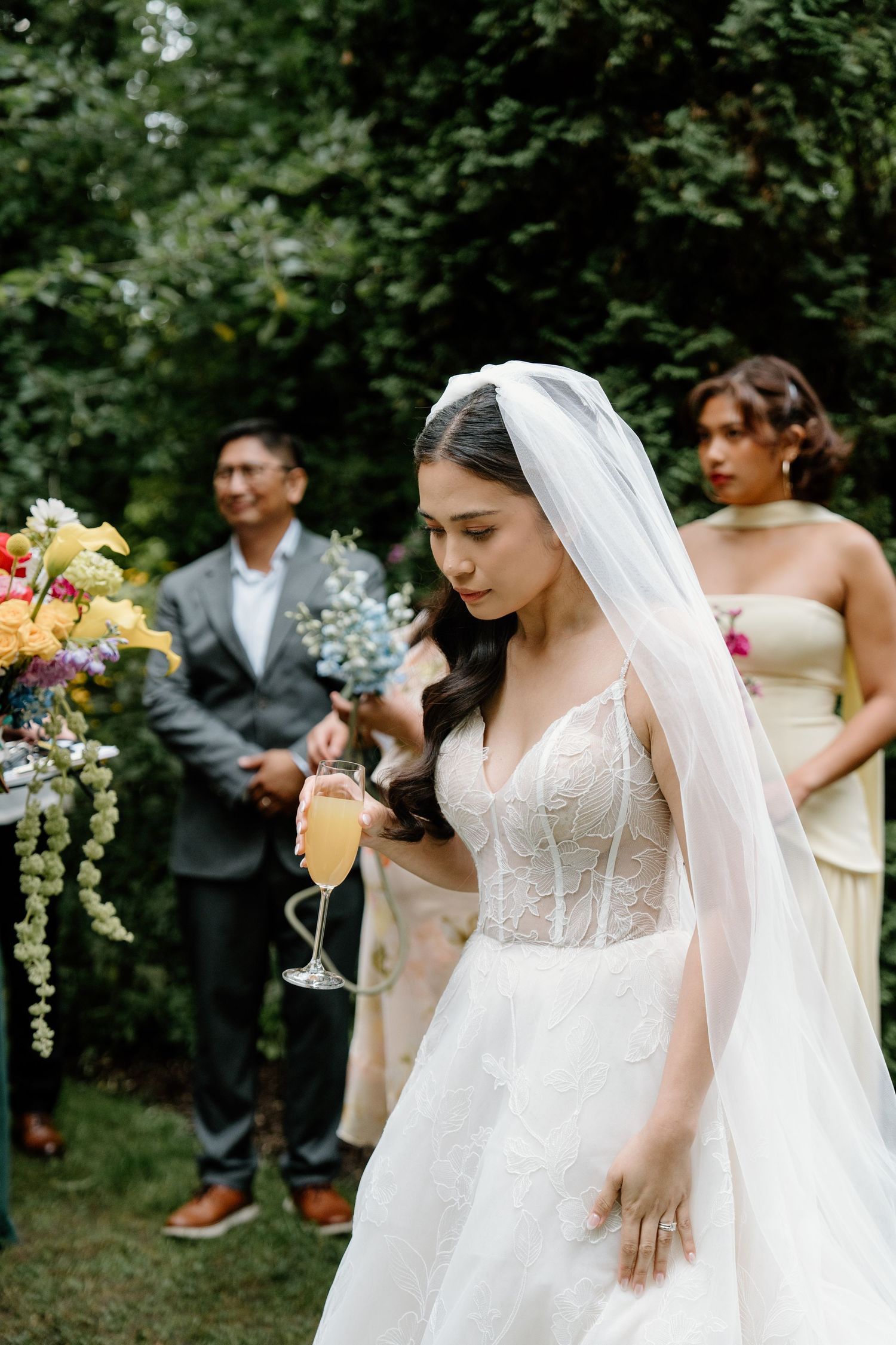 Wedding party gathered outdoors with bridesmaids in blush dresses and bride in white gown with flowing veil.