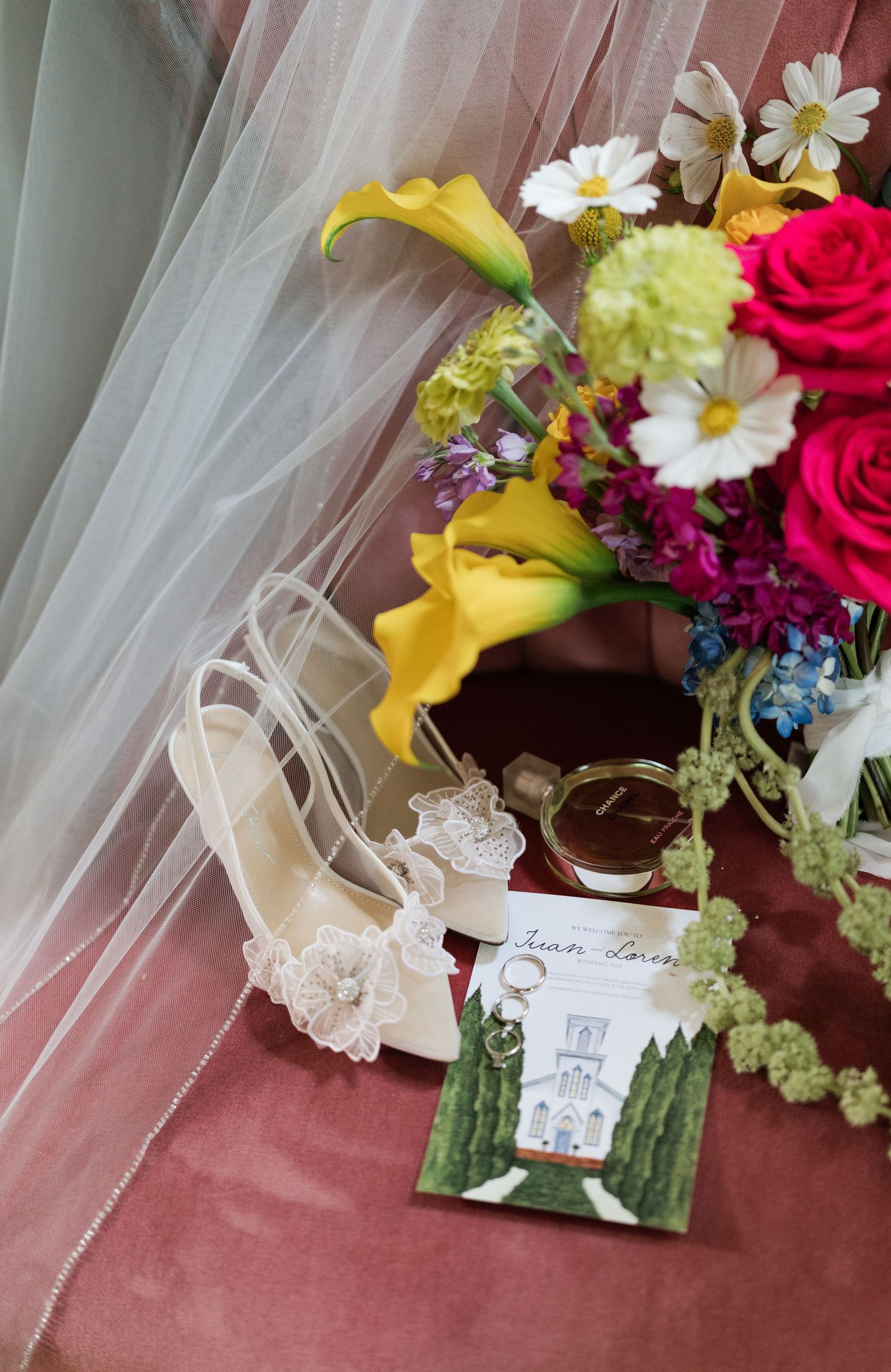 Wedding details including colorful flowers, delicate lace shoes, and invitation cards arranged on pink fabric.