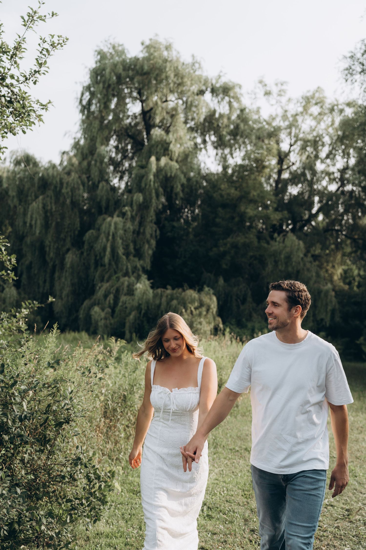 A couple in casual attire shares a joyful moment together while walking through a lush green meadow on a sunny day.