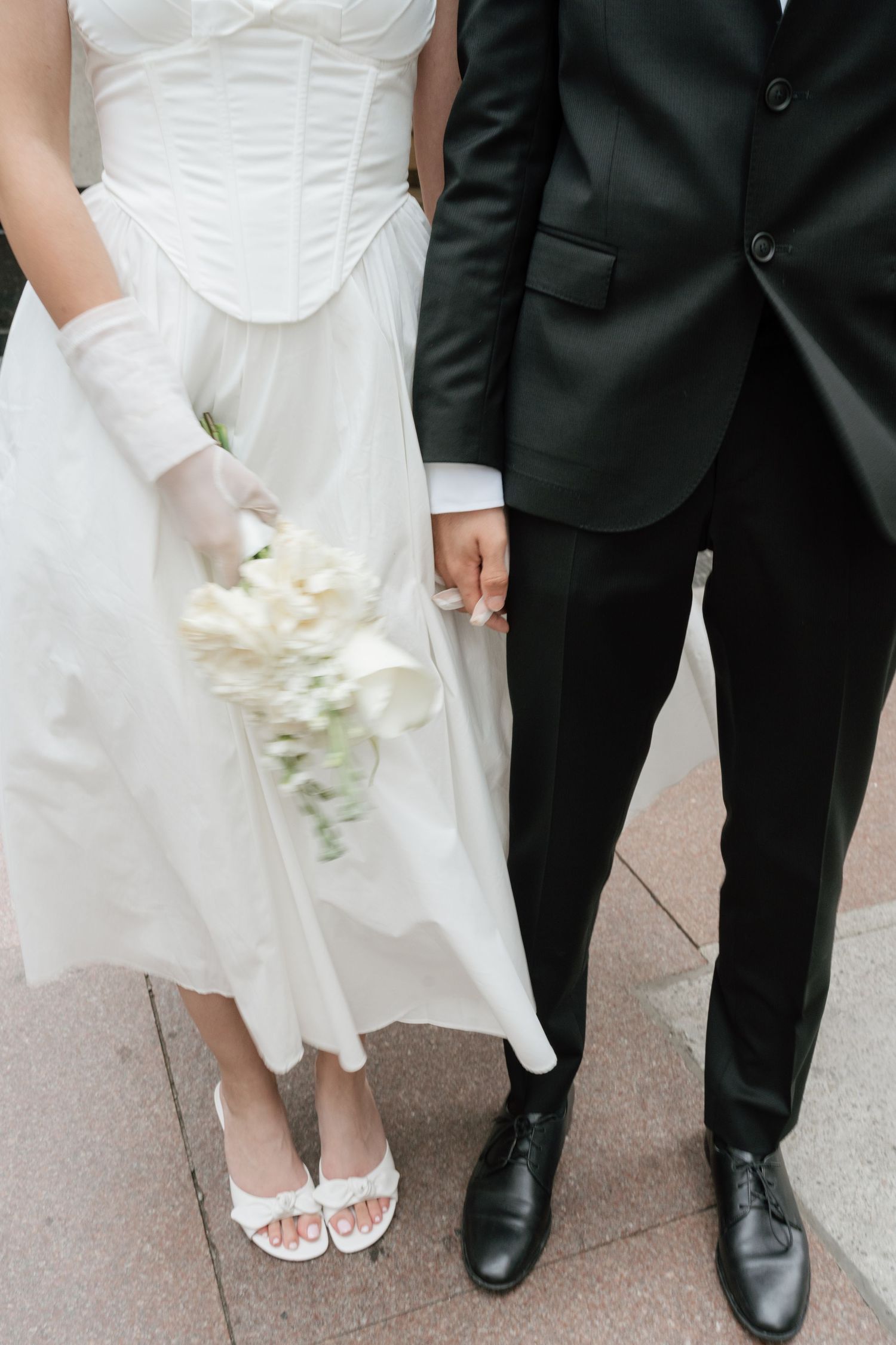 A bride in a white dress and groom in black suit standing together holding hands, with white bouquet and formal shoes.