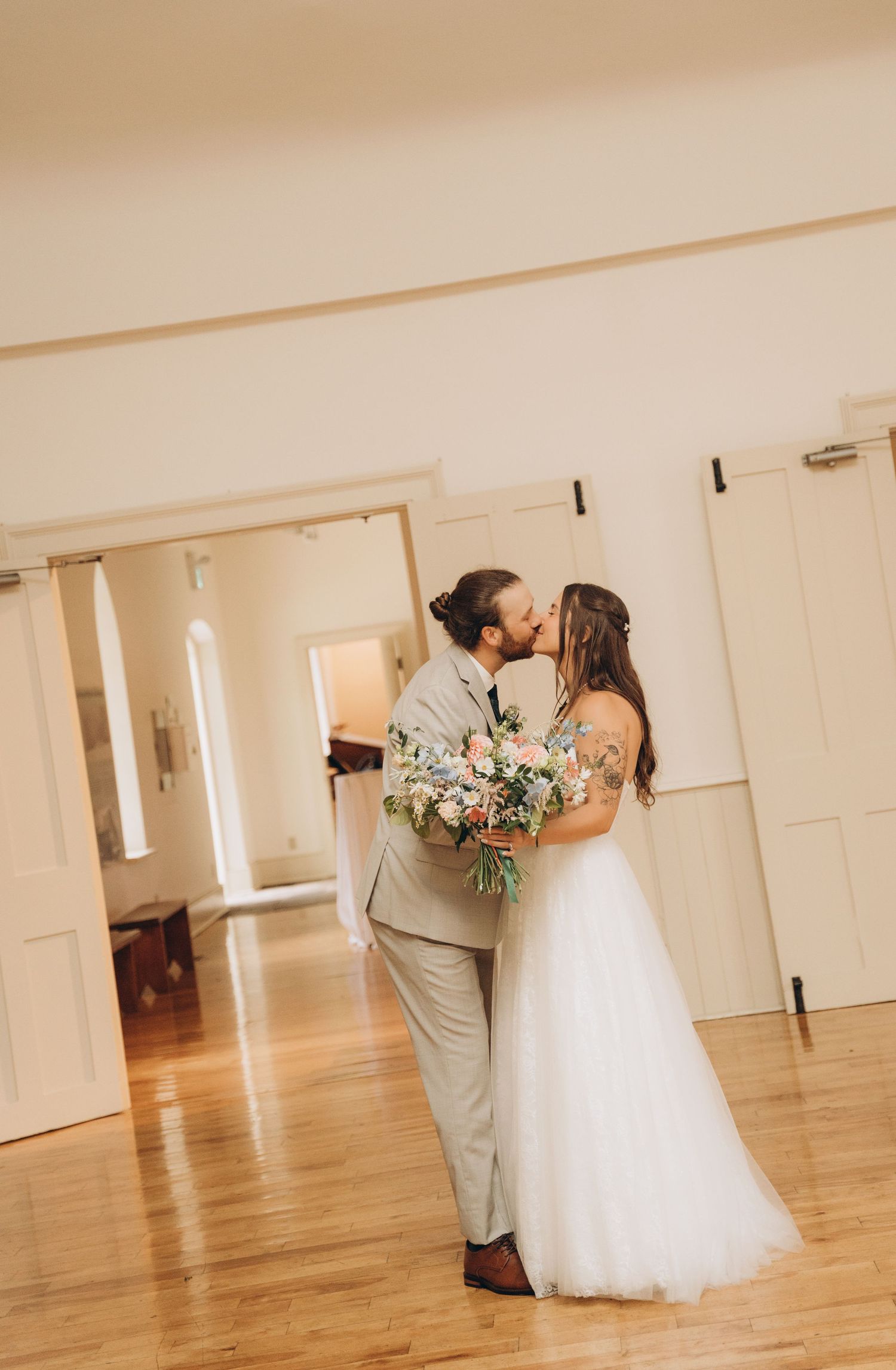A romantic wedding couple shares a tender kiss in a bright, minimalist indoor venue with hardwood floors.