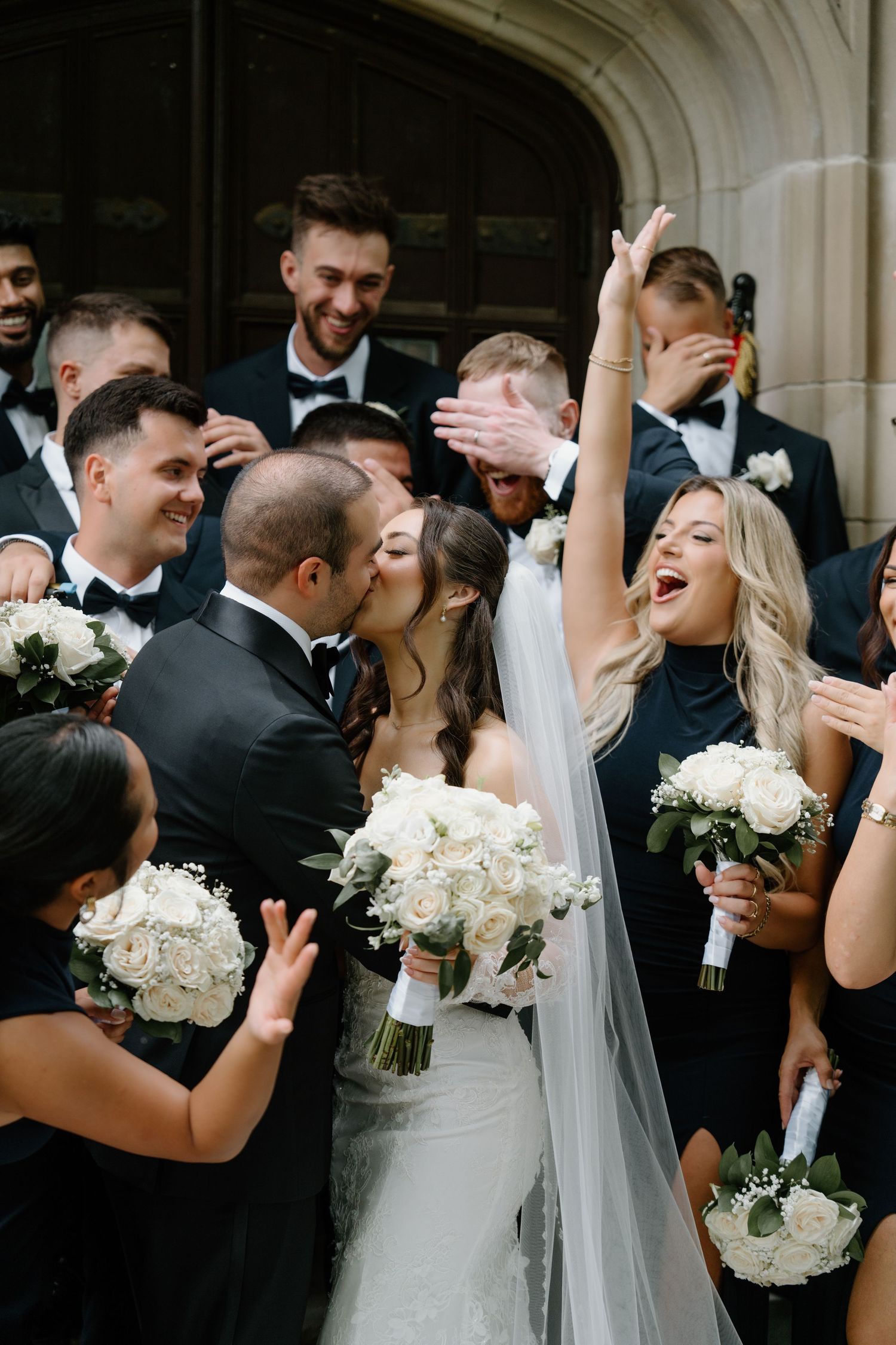 Wedding party celebrates joyfully with white bouquets in formal black and white attire at elegant venue.