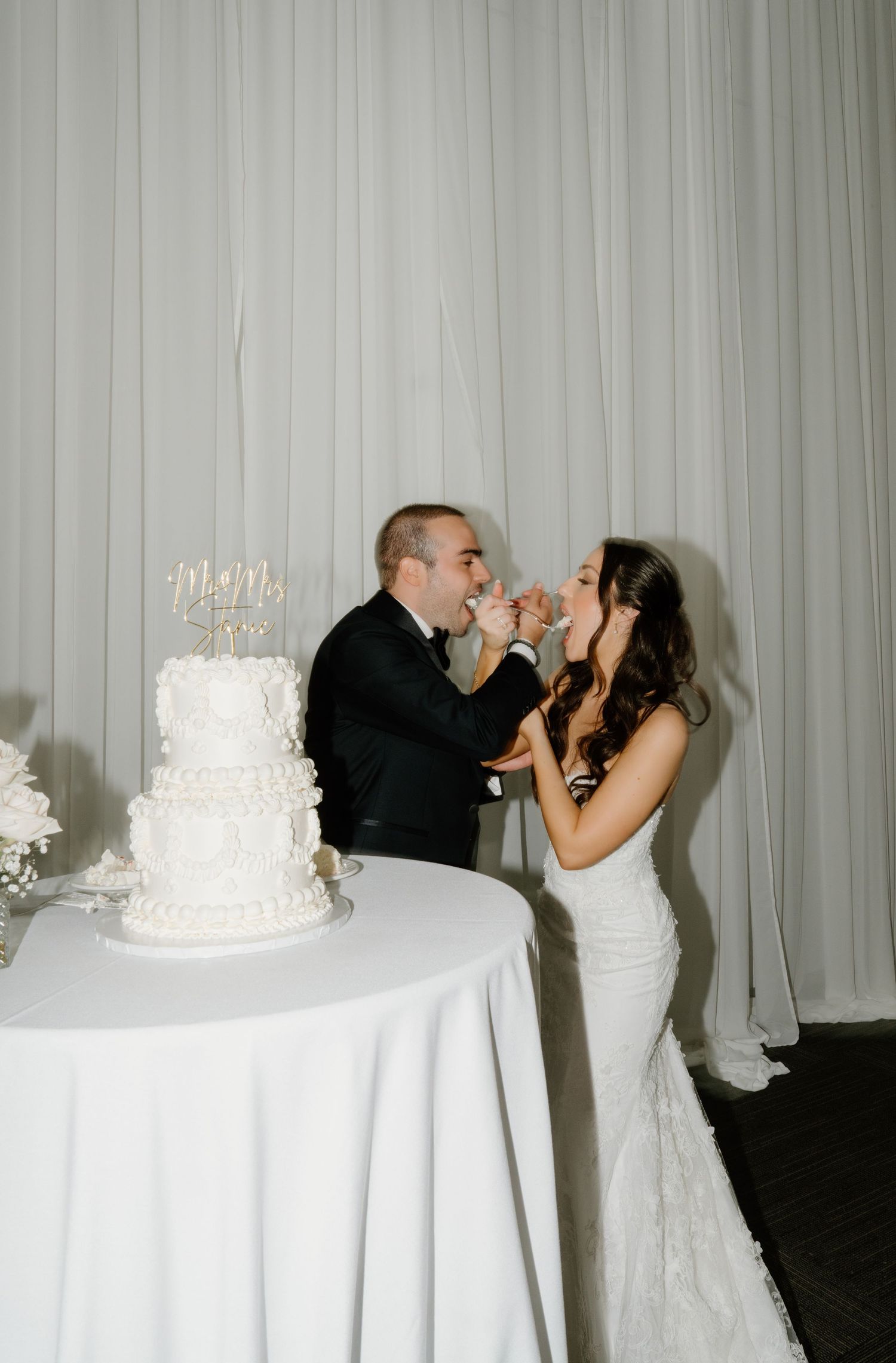 Bride and groom share a romantic moment during their wedding cake cutting ceremony with white decor and curtains.