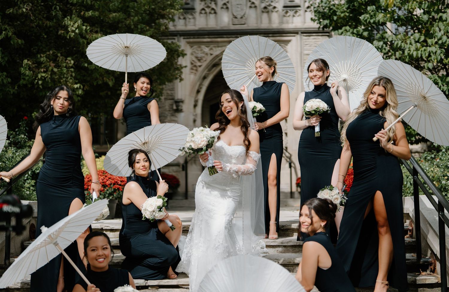 Bridal party in navy blue dresses poses with white parasols on stone steps during outdoor wedding celebration.