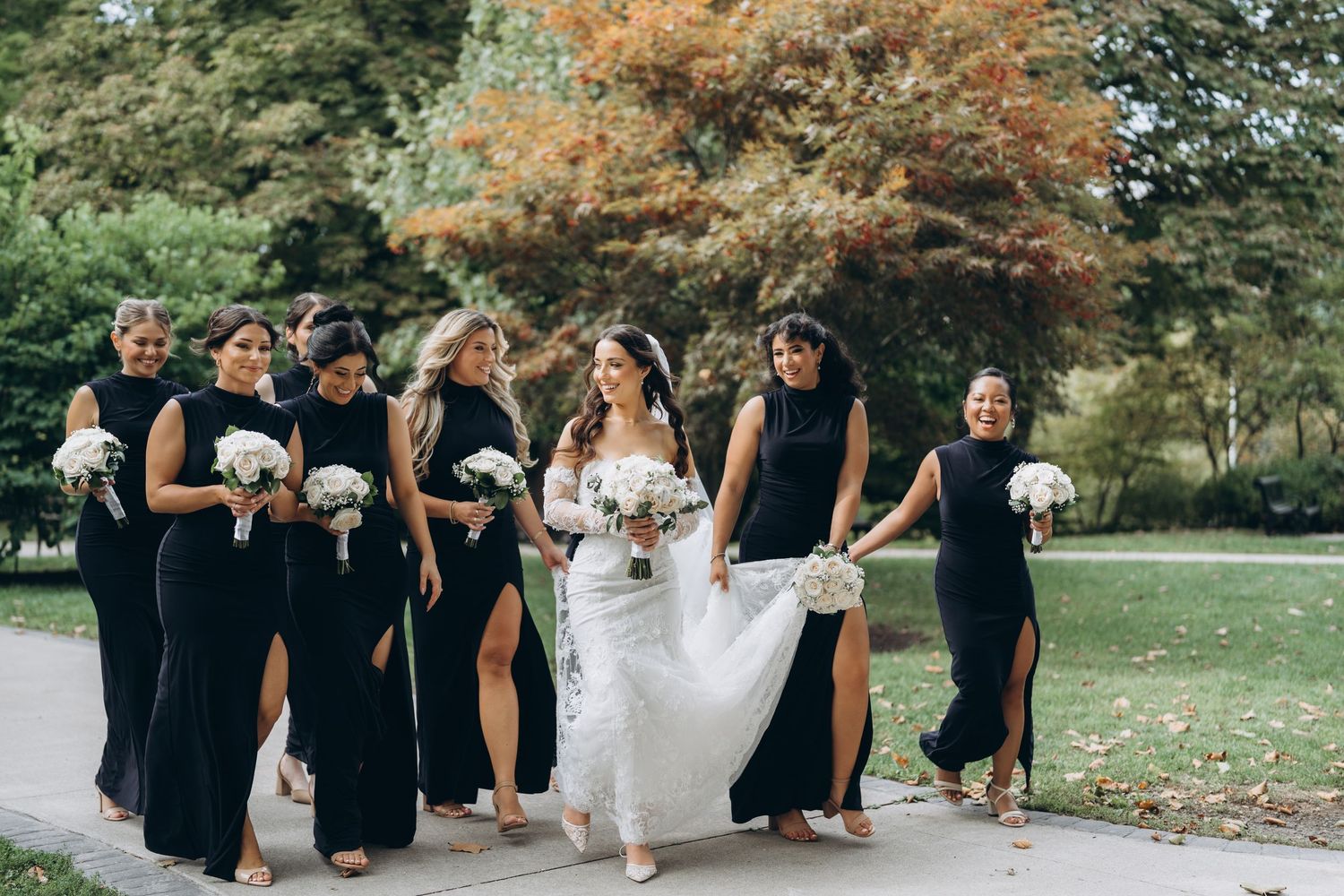 Wedding party in black dresses lifting white wedding gown in autumn garden setting.