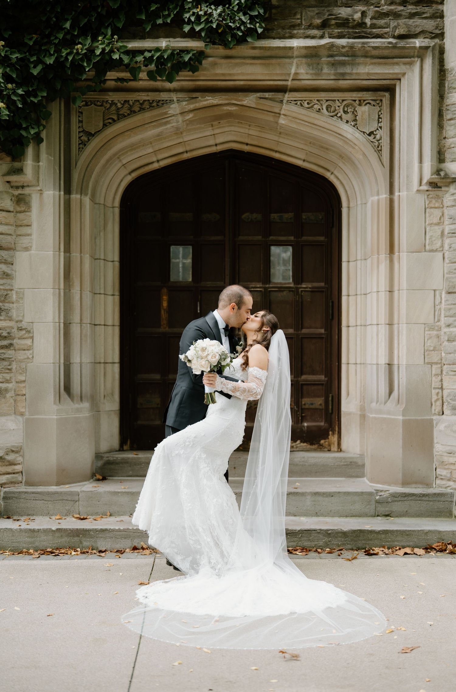 A romantic wedding couple shares a kiss in front of a grand stone church entrance with elegant archways.