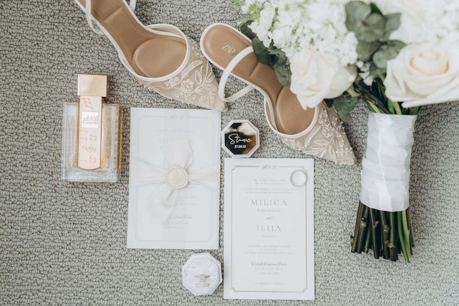 Wedding details flatlay with white shoes, invitations, perfume bottle, and rose bouquet on grey carpet.