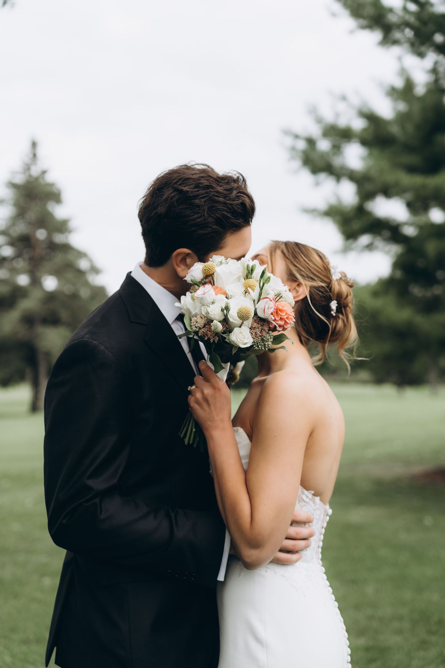 Newlywed couple shares a romantic moment behind bouquet of white and peach roses in outdoor garden setting.