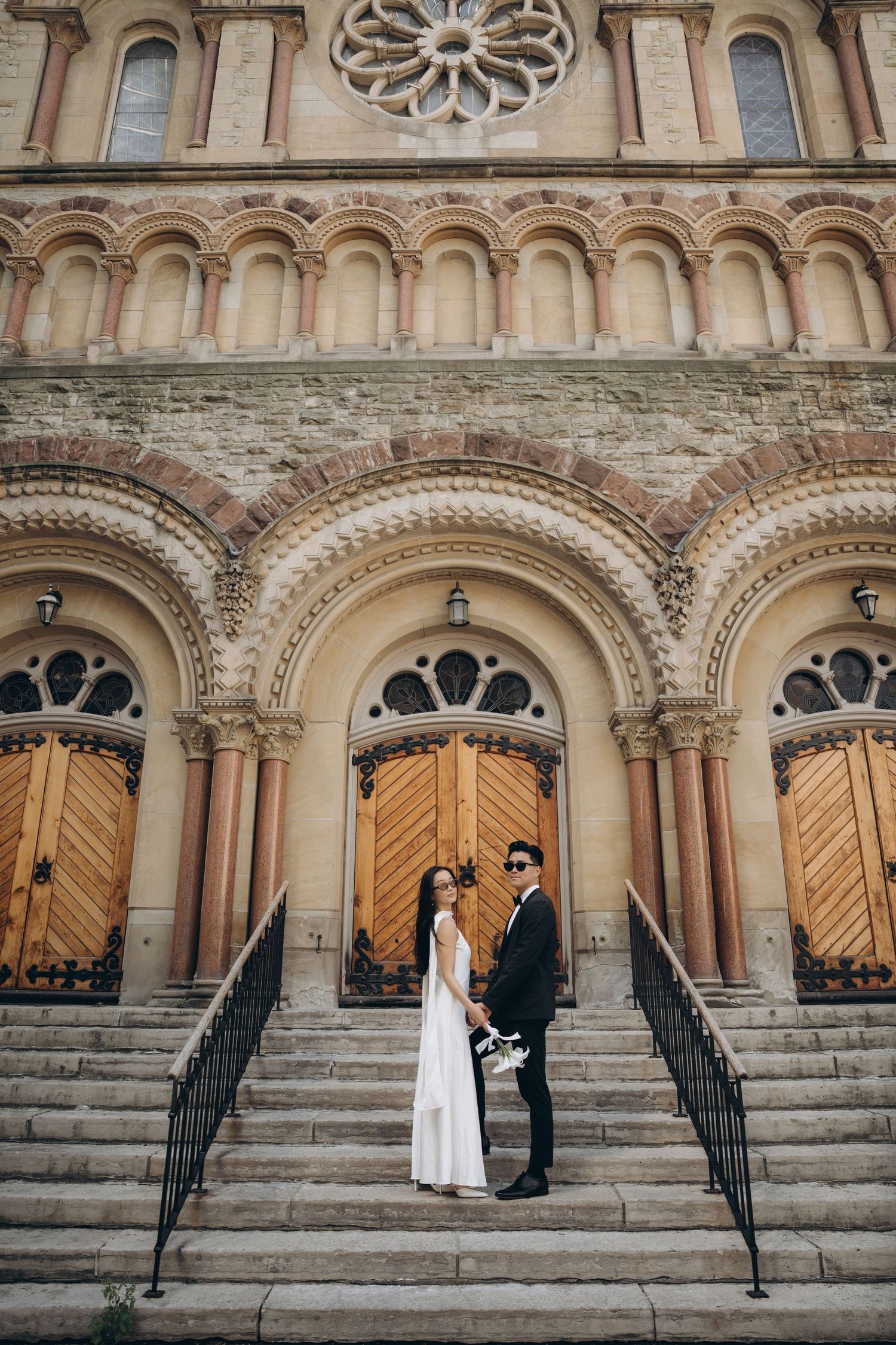 A couple stands on grand stone steps in front of an ornate church facade with arched doorways and architectural details.