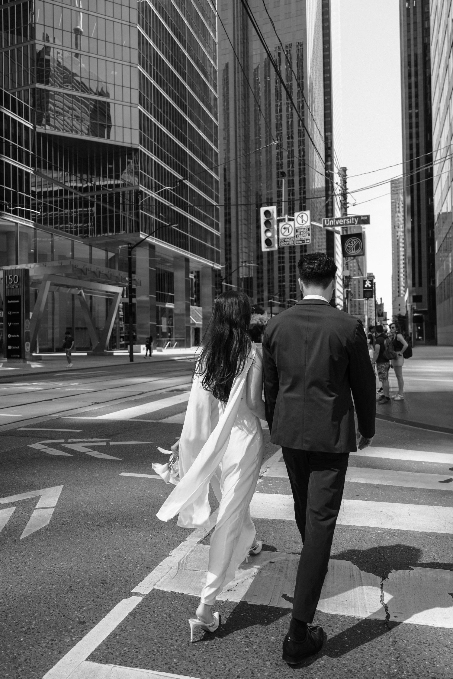 A couple in wedding attire walks across an urban intersection with modern glass buildings in the background.