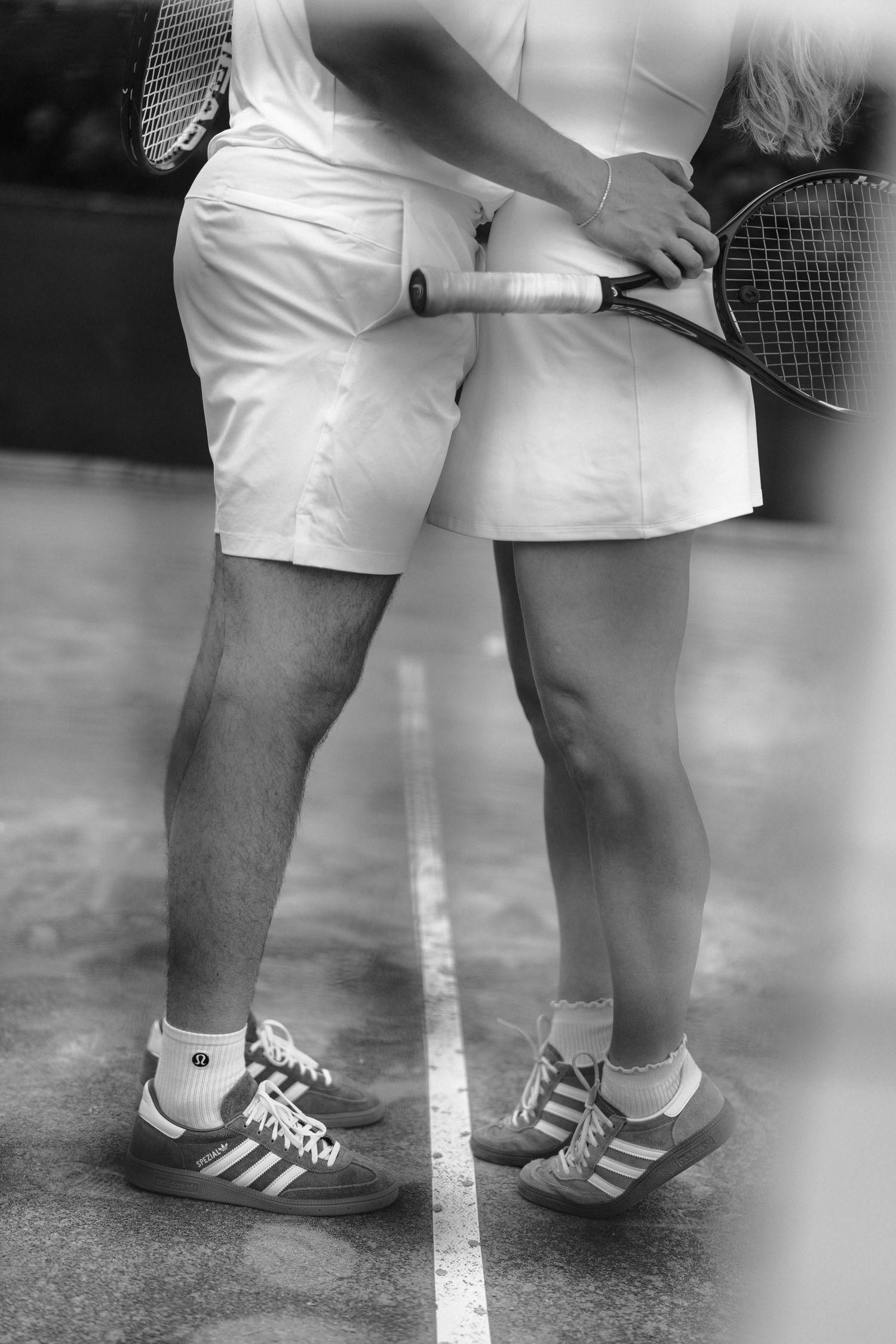 Black and white photo of tennis players' legs wearing white shorts and Adidas shoes standing on a court line.